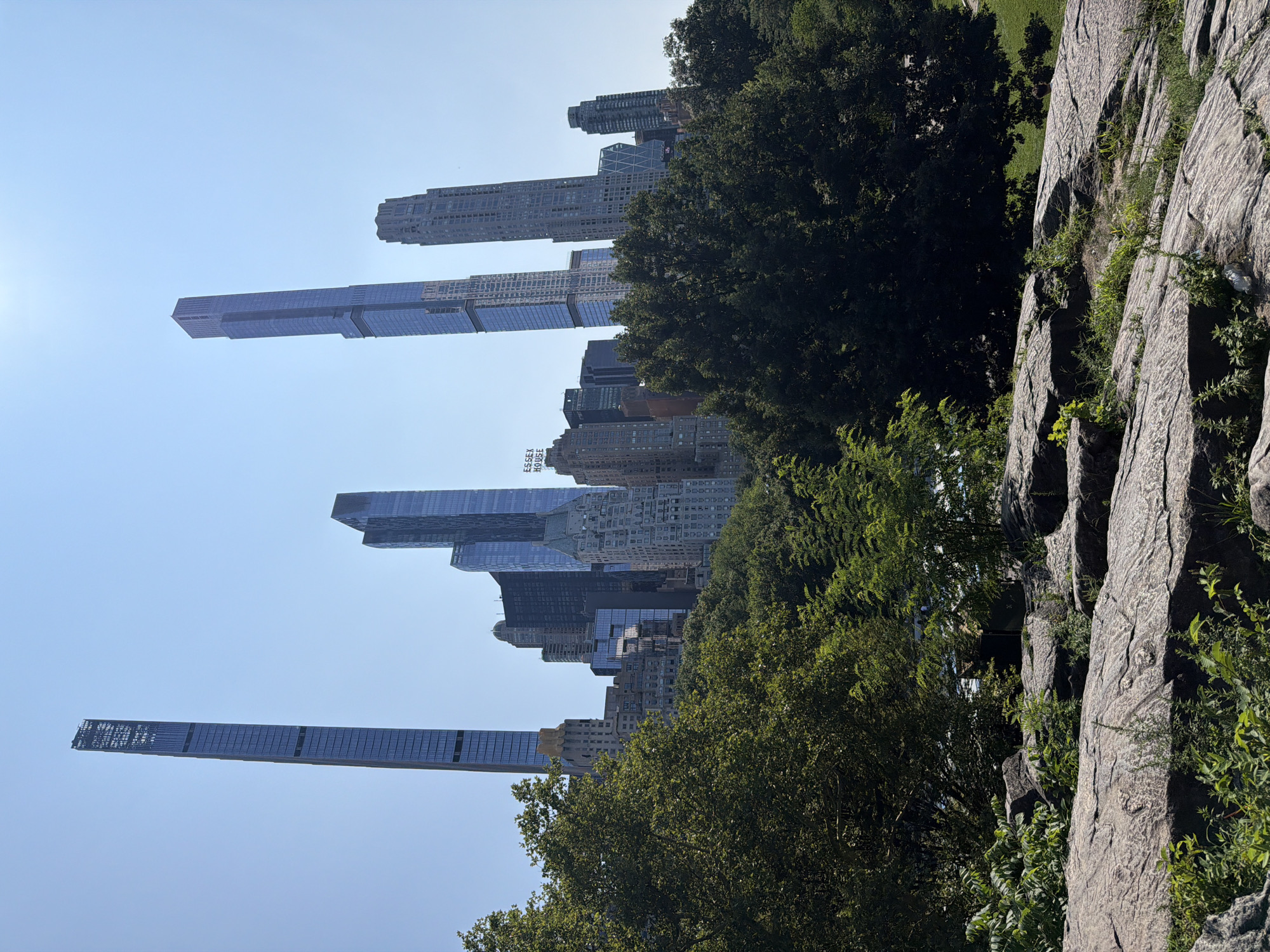 The image depicts a view of the New York City skyline as seen from Central Park. In the foreground, large, weathered rocks with sparse green vegetation are visible. Behind the rocks, a dense canopy of lush green trees obscures the lower portions of the cityscape. Towering above the trees are numerous skyscrapers, prominently featuring modern glass and steel designs. The tallest and most prominent buildings include what appear to be Central Park Tower and 111 West 57th Street, characterized by their extreme height and slender forms. Other notable structures, such as the Essex House hotel with its distinctive signage, are also discernible within the cluster of buildings. The sky is a clear, bright blue, indicating a sunny day. The overall scene contrasts the natural landscape of the park with the impressive architectural achievements of the city.