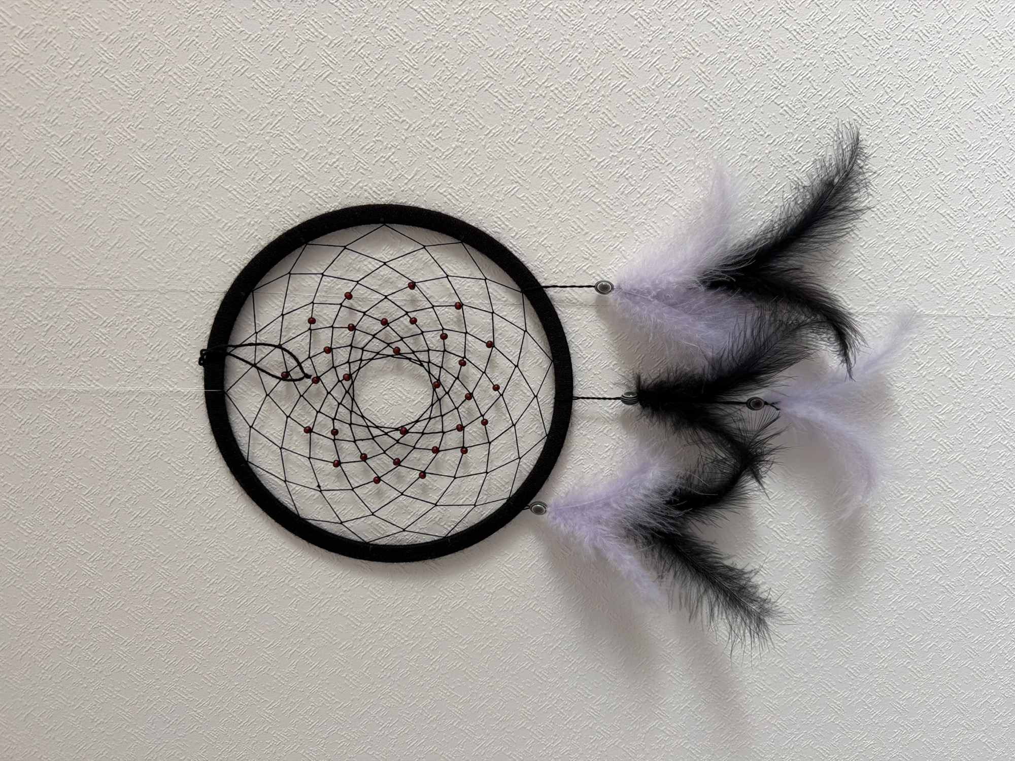 The image shows a dreamcatcher hanging on a white textured wall. The dreamcatcher has a black circular frame with a web of black string inside, dotted with small red beads. Attached to the right side of the frame are three sets of black and white feathers. The image gives no indication of the location being Moscow, Russia.