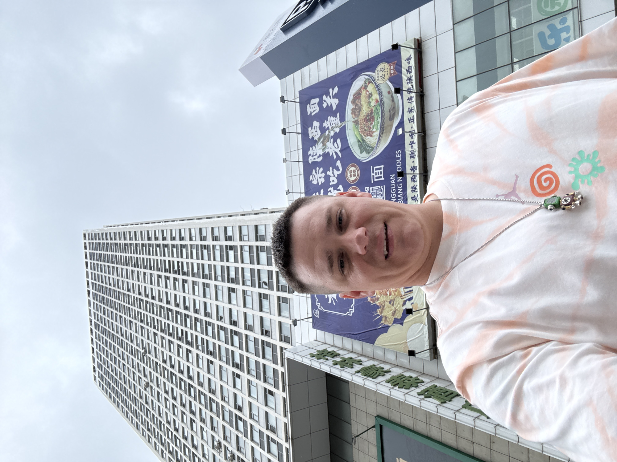 A man is taking a selfie outdoors in front of a tall, modern building in Qinhuangdao, China. The building has a grid of numerous windows. To the right of the building, a large, blue billboard displays an image of a bowl of noodles and text in Chinese characters. The man is wearing a tie-dye t-shirt with peach and white colors and a silver chain with a pendant. The sky is overcast.