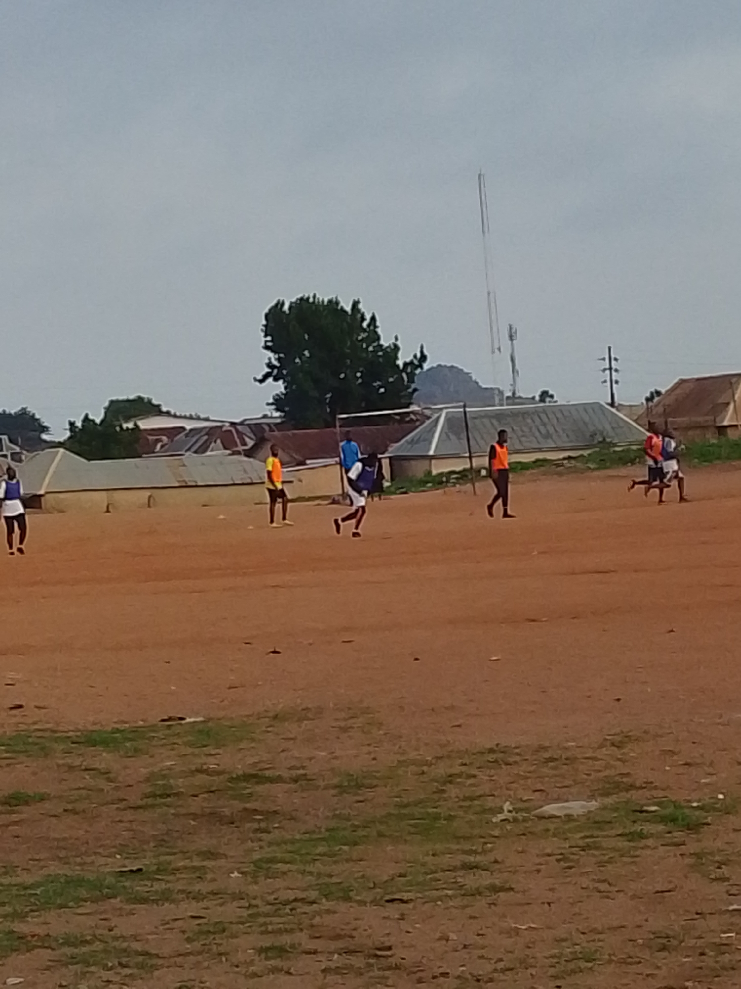 The image depicts a dusty, open field in Bukuru, Nigeria, where several people are engaged in some form of physical activity. The ground is predominantly reddish-brown with sparse patches of green vegetation. Buildings with corrugated metal roofs line the perimeter of the field, and a tree is near the center-background. In the background, an antennae. The sky is overcast. The people are wearing different colored vests and performing activities.