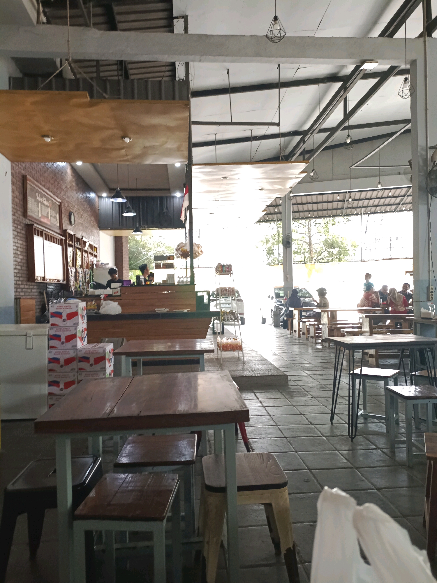 The image depicts an open-air food court or dining area, likely in Made, Indonesia. In the foreground, several wooden tables with metal legs are arranged, some with empty stools. To the left, stacks of boxes with red and white branding are visible. Further back, a counter area is staffed by at least two individuals wearing dark shirts. Beyond the counter, the space opens up to an outdoor area with more tables and people seated, suggesting a public gathering space. Cars are parked in the background, and a tree is visible, indicating an outdoor or semi-outdoor environment. The architecture features exposed beams and a corrugated metal roof. The overall impression is of a casual dining establishment or a community space.