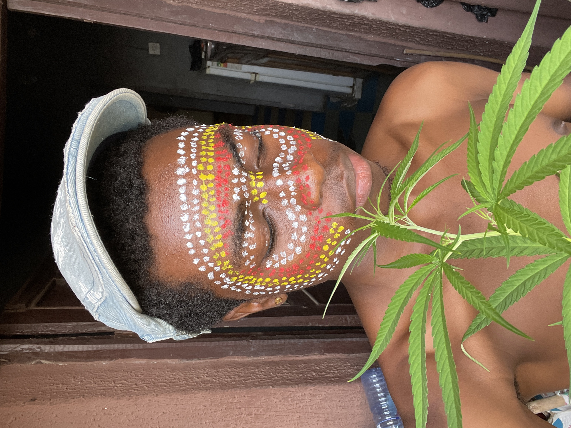 A man with brown skin, wearing a light-blue denim hat, is positioned in front of a window frame. His face is decorated with patterns of white, yellow, and red dots. He is topless and a cannabis plant covers part of his body. A water bottle is in the lower center of the frame. The image was taken in Nkpor, Nigeria.