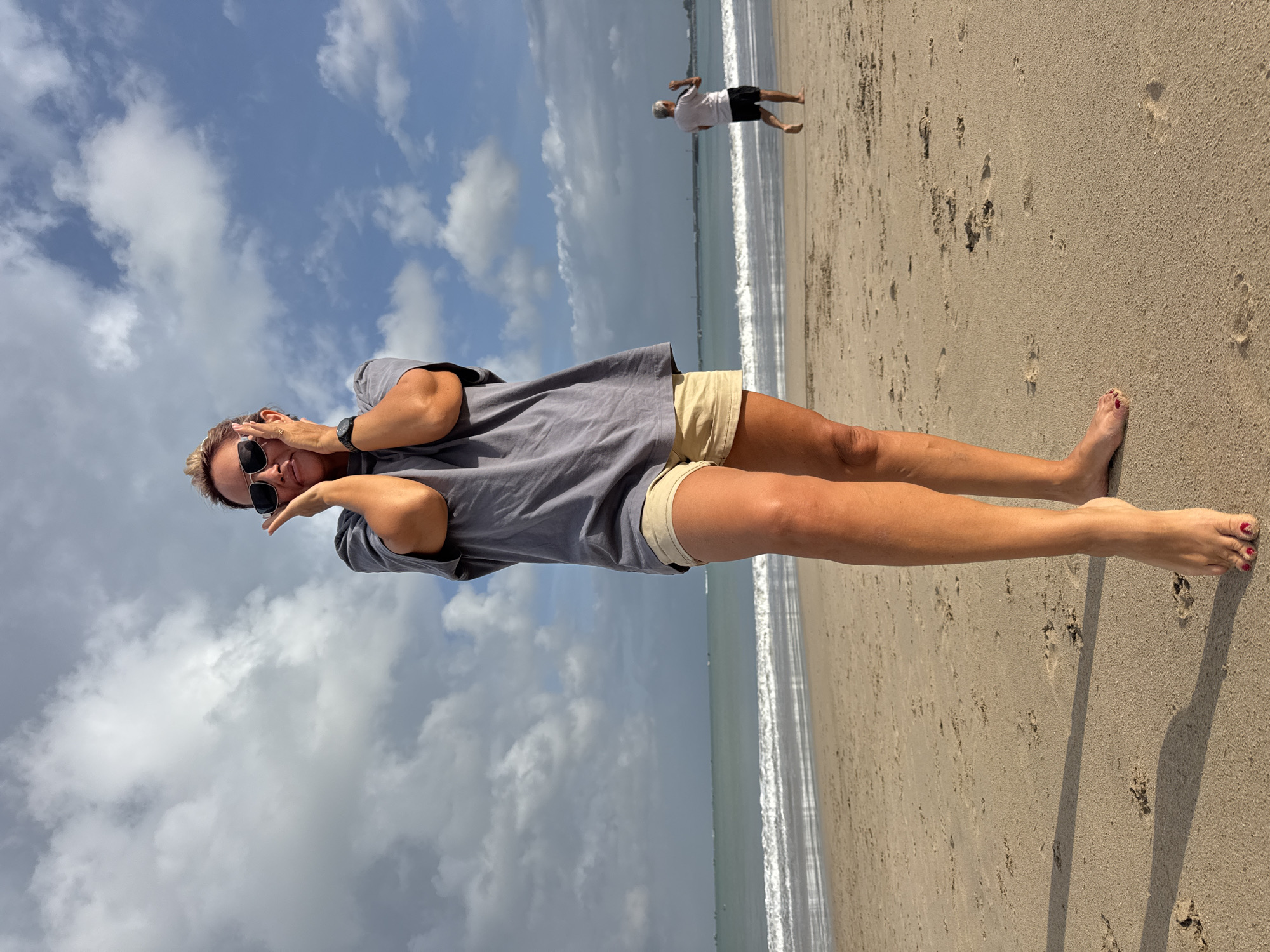 A woman with short, blonde hair and sunglasses stands on a sandy beach, her hands pressed against her cheeks. She wears a grey t-shirt and beige shorts. The ocean with small waves is in the background, and a man in a white shirt and black shorts walks in the distance. The sky is partly cloudy. The image is taken in Kuta, Bali, Indonesia.