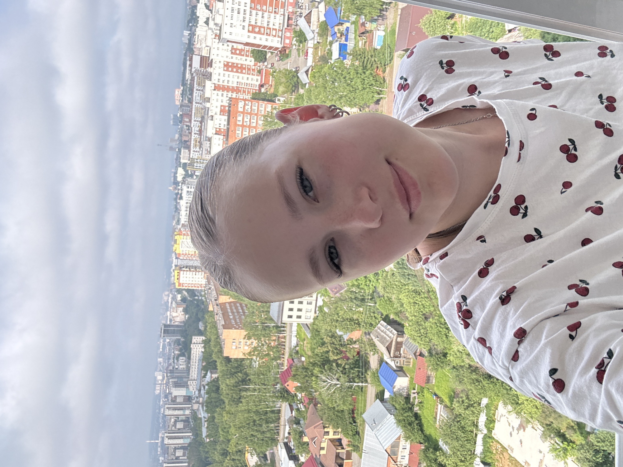 The image is a selfie of a young girl taken from a high vantage point, likely a balcony or window, overlooking a city. The girl is positioned in the foreground, with her face tilted towards the camera and her body at an angle. She has blonde hair, blue eyes, and is wearing a white t-shirt with a repeating pattern of red cherries. She has a slight smile, and her left ear has multiple piercings.
The background is a sprawling cityscape with a mix of tall apartment buildings and smaller houses with red and blue roofs, interspersed with lush green trees. The architecture suggests an urban environment, and the presence of many trees indicates a location with significant greenery within the city. The sky is overcast and a muted grey, suggesting cloudy or potentially rainy weather. The overall lighting is soft and diffused, which could indicate it is daytime, but not necessarily sunny.
The setting appears to be a residential area within a city. The perspective suggests the photo was taken from several stories up. There is no visible text in the image. The photo captures a moment of casual self-expression by the girl, set against the backdrop of the urban landscape. The orientation of the photo is sideways, as if taken while holding the phone vertically.