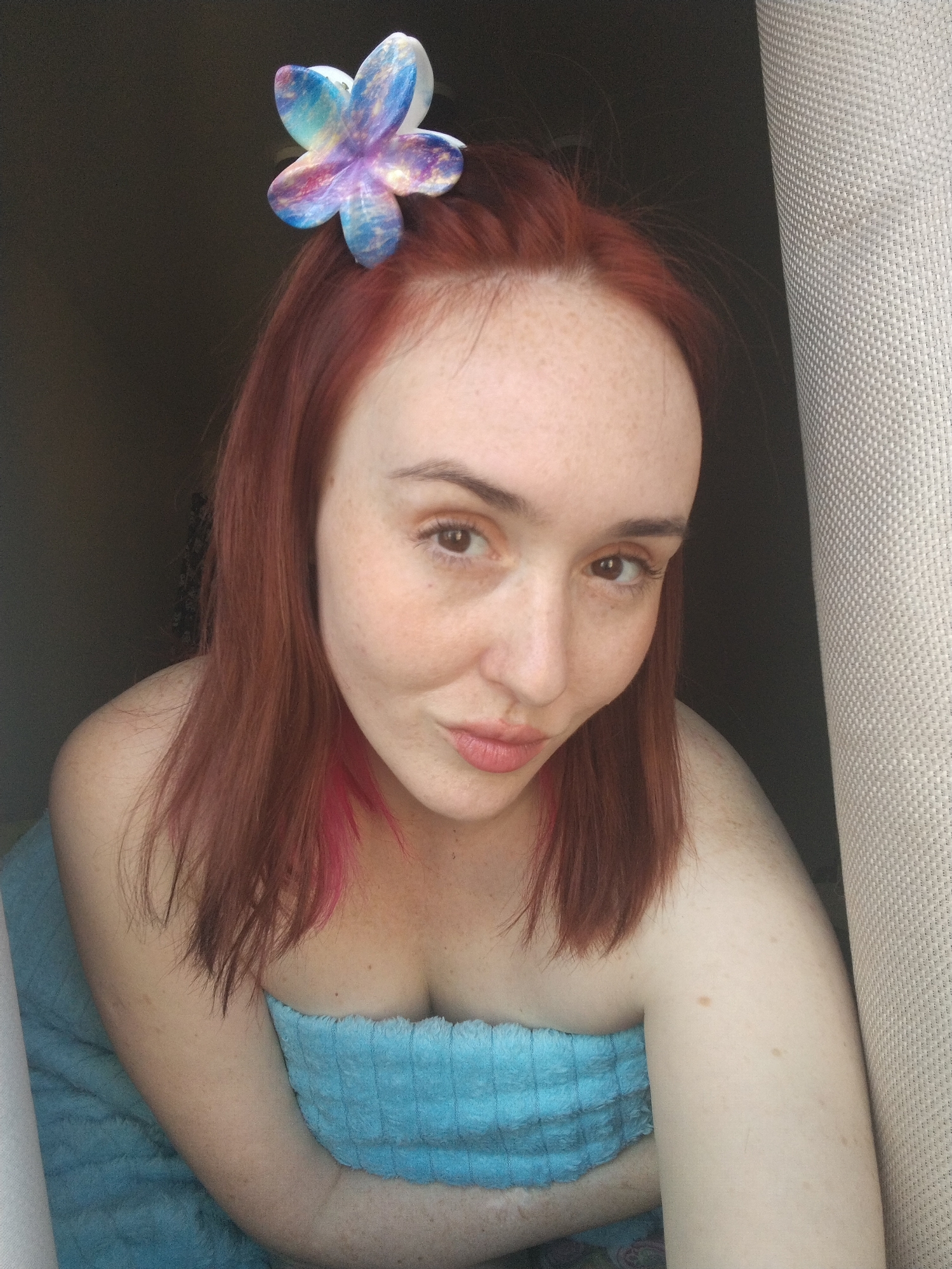 A selfie depicts a woman with fair skin and red hair, with a blue and purple flower hair accessory on the left side of her head. She has freckles across her face and is wearing a blue towel. The background includes a dark shadowed area and a light gray textured surface, possibly a curtain or wall. The location is unspecified and there is no information indicating the location is Izhevsk, Russia.
