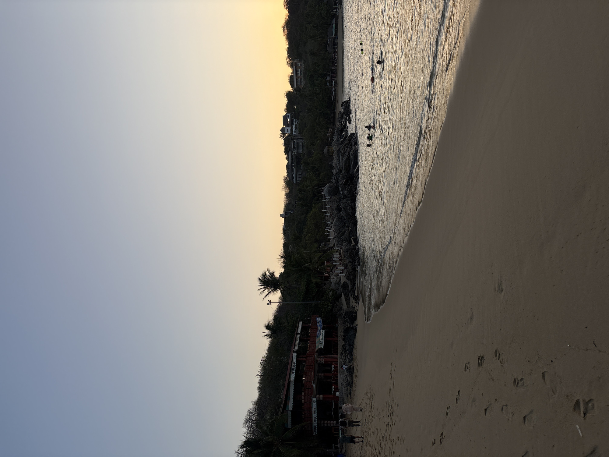 The image depicts Zicatela Beach in Puerto Escondido, Mexico, at sunset. The sky transitions from blue to orange. The scene includes the coastline where the ocean meets the sandy beach. There are a few people swimming in the ocean. There are a few people standing on the beach near a restaurant. A hillside with vegetation and a building is visible behind the beach. Footprints are visible in the sand on the right side of the image.