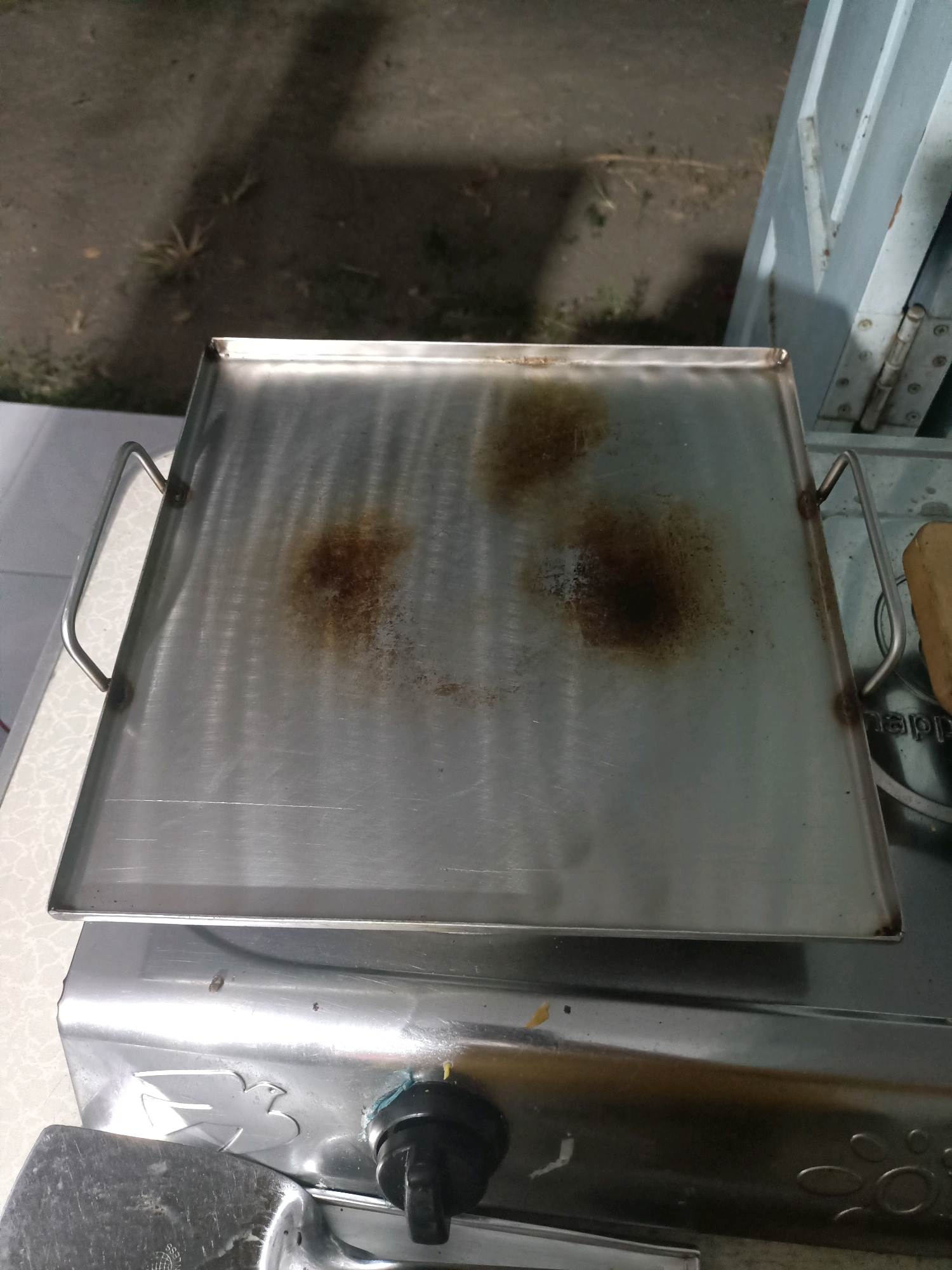 A stainless steel griddle with two handles sits atop a gas stove. The griddle has several burnt-on stains. The stove is metallic with a decorative embossed pattern and a black control knob. In the background, a light-colored countertop is visible, as is a gray, hinged metal structure that appears to be a gate or door. The ground beyond the countertop is visible and appears to be concrete or asphalt with some scattered debris. The scene is dimly lit, suggesting it may be nighttime or indoors with artificial lighting. The location is identified as Badas, Indonesia.