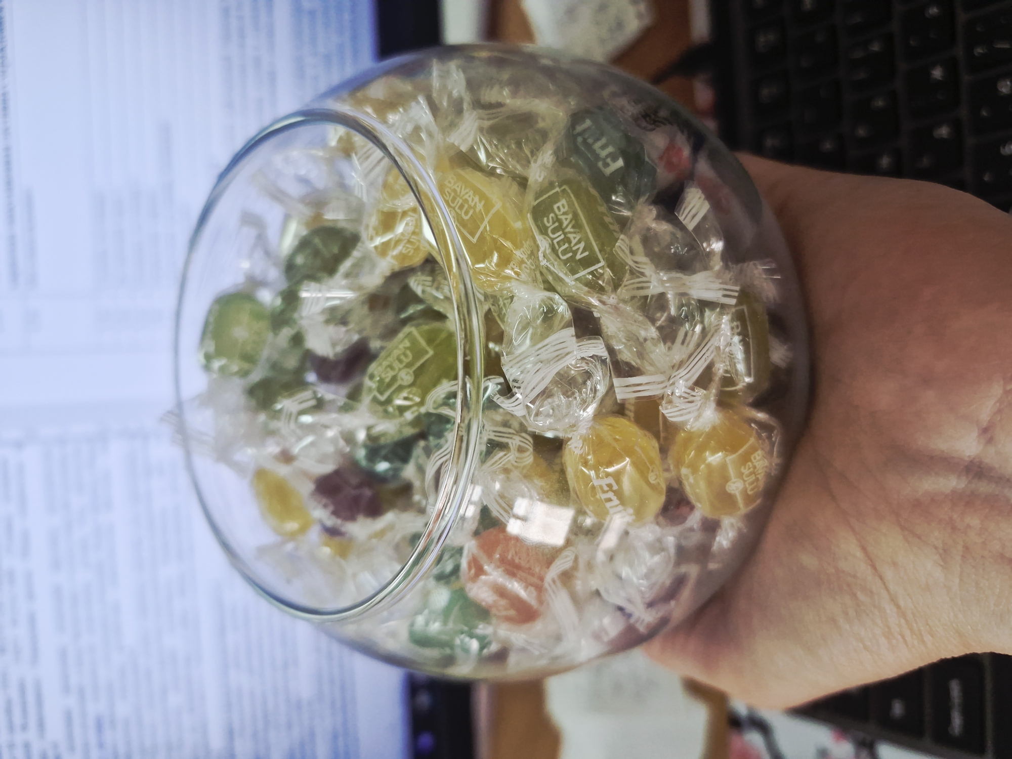 A close-up image displays a human hand holding a clear, spherical glass bowl filled with individually wrapped hard candies. The hand, with visible skin texture, supports the bowl from beneath. Inside, numerous candies in vibrant colors such as yellow, light green, dark green, orange, and purple are visible through their clear plastic wrappers. Many of the wrappers have distinctive white twisted ends.
Text on several yellow and green candy wrappers clearly reads "BAYAN SULU". Other wrappers also show partial text like "Emin" or "Emli". The background is softly blurred: a white document with black text is visible to the left of the bowl, and a dark-colored keyboard is present to the right. This suggests an indoor setting, likely an office or desk environment. The scene is brightly and evenly lit. The image captures the act of holding or presenting the candy-filled bowl.