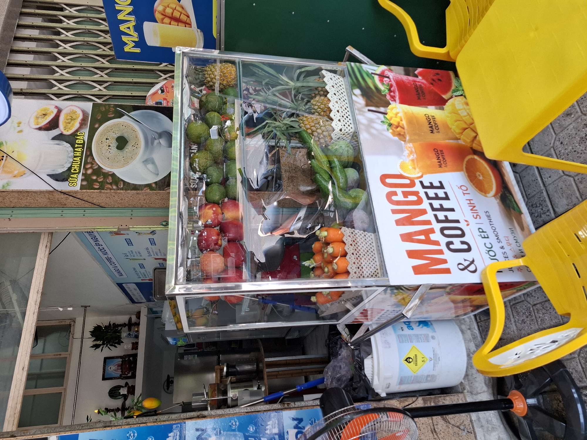 The image captures a vibrant street-side juice and coffee stand in Nha Trang Ward, Vietnam. In the foreground, a bright yellow plastic chair and table are set up, suggesting a seating area for customers. Behind the furniture, a refrigerated display case showcases an assortment of fresh fruits, including apples, avocados, pineapples, and a basket of carrots. A sign above the display advertises "MANGO & COFFEE" in large, bold lettering, along with images of colorful fruit smoothies and an orange slice.
Further back, various items related to the business are visible. A large white bucket with hazard symbols is placed near a black fan with an orange motor. To the left, a blue and white sign displays text in Vietnamese and English, possibly indicating menu items or pricing. The background reveals a glimpse of the shop's interior, with shelves displaying more items, and a scooter parked within. The overall scene is bustling and colorful, indicative of a typical Vietnamese street food or beverage vendor.