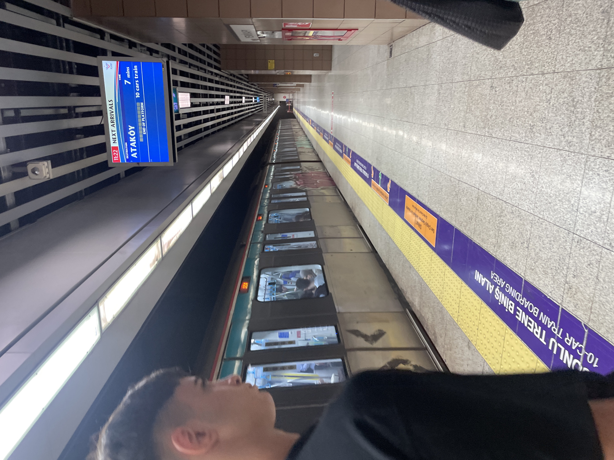 The image depicts a subway station platform in Istanbul, Turkey. A train is stopped at the platform, and passengers are visible inside the train cars. A digital display board shows "NEXT ARRIVALS" with "ATAKÖY" listed as the next destination, indicating a 7-minute wait for a 10-car train. The platform floor is tiled and has yellow tactile paving along the edge, with purple boarding area markers indicating "10-CAR TRAIN BOARDING AREA." In the foreground, a person is partially visible on the left, facing the train. The station has a modern design with a suspended ceiling featuring lights and structural elements. Signs in Turkish are visible on the walls and pillars. The overall scene suggests a busy public transportation hub.