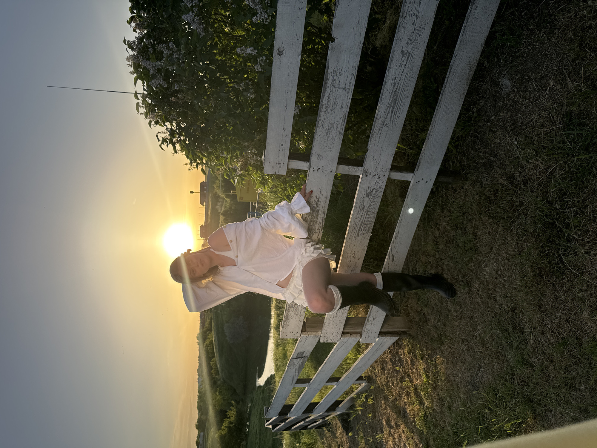 A person is reclining on a dilapidated white wooden fence in a rural setting, likely near Усть-Курдюм, Russia, as indicated by the prompt. The person is wearing a white, ruffled outfit and dark, knee-high boots. The sun is setting or rising in the background, creating a strong backlighting effect. The environment includes grassy areas, trees, and a body of water in the distance.