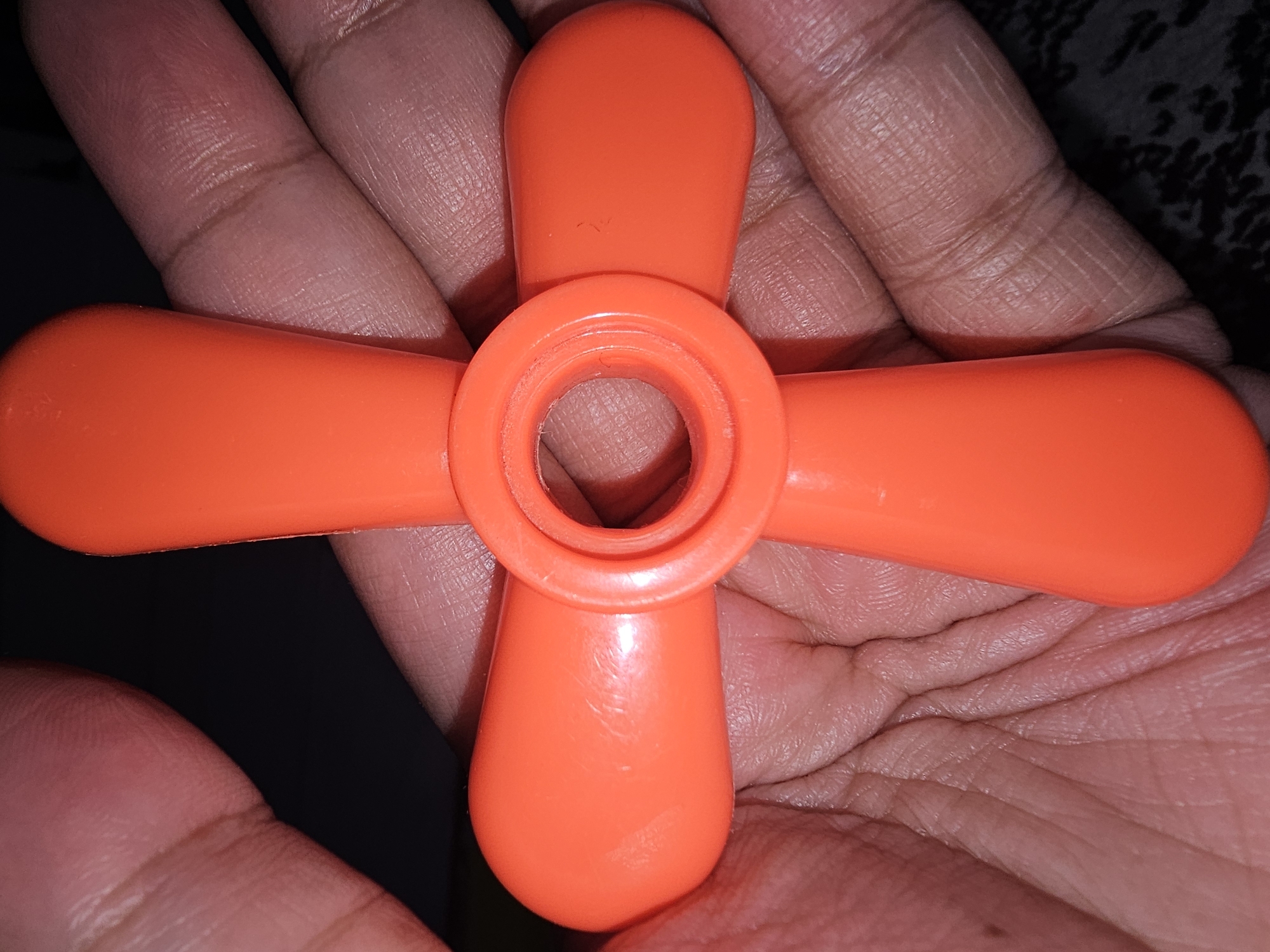 An orange, four-bladed propeller-like toy is held in a person's hand, with fingers and palm visible beneath it. The toy has a circular opening in the center. The background is dark, with some textured fabric visible at the bottom right. This image was taken in Lamongan, Indonesia.