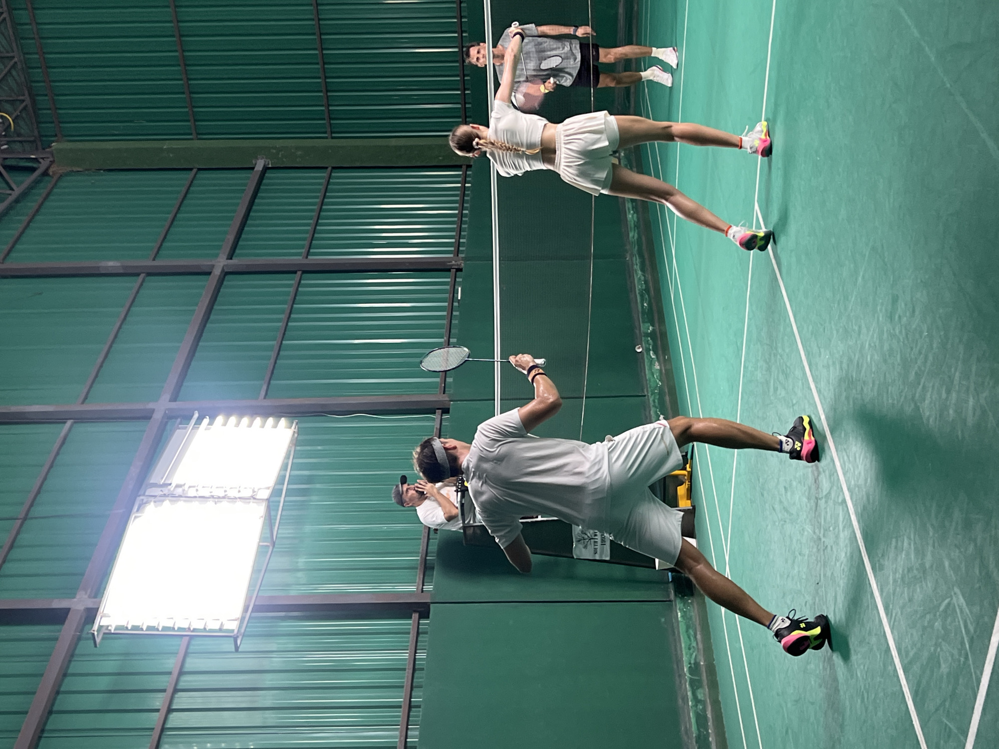 The image shows a badminton game in progress in Unknown, Unknown. Four people are present. A man in white sportswear is in the foreground, holding a badminton racket in the air, presumably about to hit the shuttlecock. Behind him are two other men, one wearing a visor and the other wearing grey sportswear. In the background, a woman in white sportswear is standing opposite the man with the racket, ready to play. The court is marked with white lines and the walls are green. A bright light fixture is mounted on the ceiling. The roof structure is visible, made of metal beams and corrugated green panels.