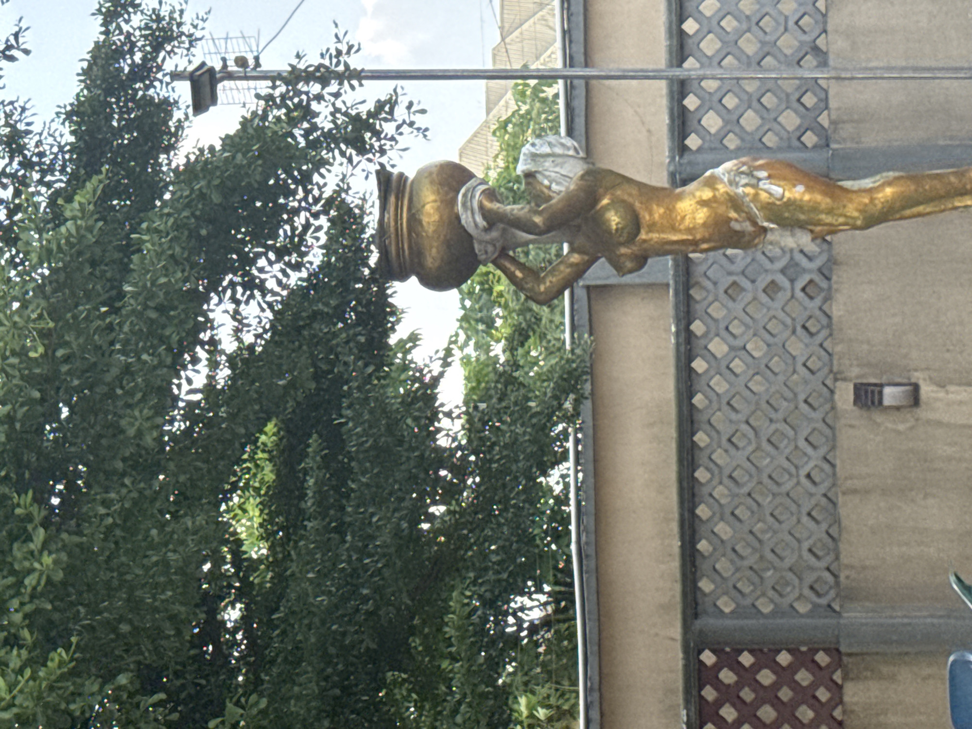 The image features a prominent golden statue of a partially nude female figure, extended horizontally from a building. The statue, rendered in a classical style, wears a white or grey head covering and holds a large, ornate golden urn or vessel with both hands. Some areas of the statue show wear, with the gold finish flaking off to reveal a lighter material underneath, notably on her head, chest, and hip.
Behind the statue and to the left, dense green foliage from trees or bushes fills a significant portion of the frame. Sunlight filters through the leaves, creating bright highlights and dappled light effects. Above the foliage, the sky is visible, appearing bright and possibly hazy. An antenna-like structure is mounted on a pole and partially obscured by the trees in the upper left corner.
The building the statue is attached to features a distinctive patterned facade. This facade displays a repeating geometric design, resembling a lattice or tessellation of squares or diamonds, primarily in shades of grey. A lower section of the building exhibits a similar pattern but in a darker, reddish-brown hue. A horizontal metal bar supports the statue and crosses the scene. A small, square light fixture or vent is also visible on the building wall to the right.
The scene depicts a daytime setting. No visible text is present in the image. The environment suggests an outdoor, possibly urban or suburban, location in Ihiagwa, Nigeria, characterized by both architectural details and natural greenery.