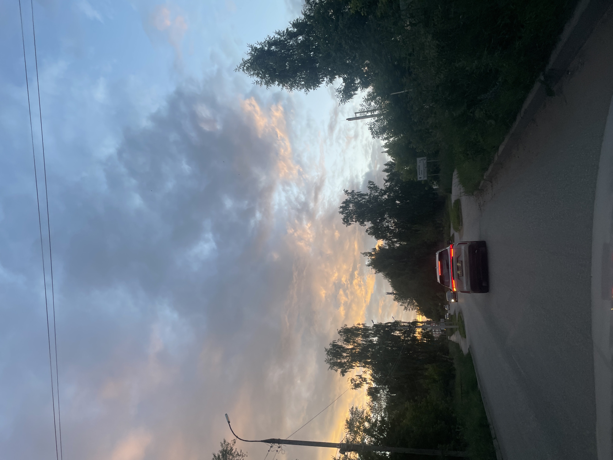The image shows a road with a car driving away from the camera. The car is silver with its brake lights illuminated. The road is paved and curves to the right. On either side of the road are trees and foliage. In the background, there is a cloudy sky with patches of pink and orange, suggesting it is either sunrise or sunset. There are power lines visible in the sky and a street lamp on the left side of the road. A street sign is visible on the right side of the road, with text that is partially legible but appears to be in Cyrillic script and likely indicates a direction or place name. The overall scene conveys a sense of a quiet, rural or suburban road at dusk or dawn.