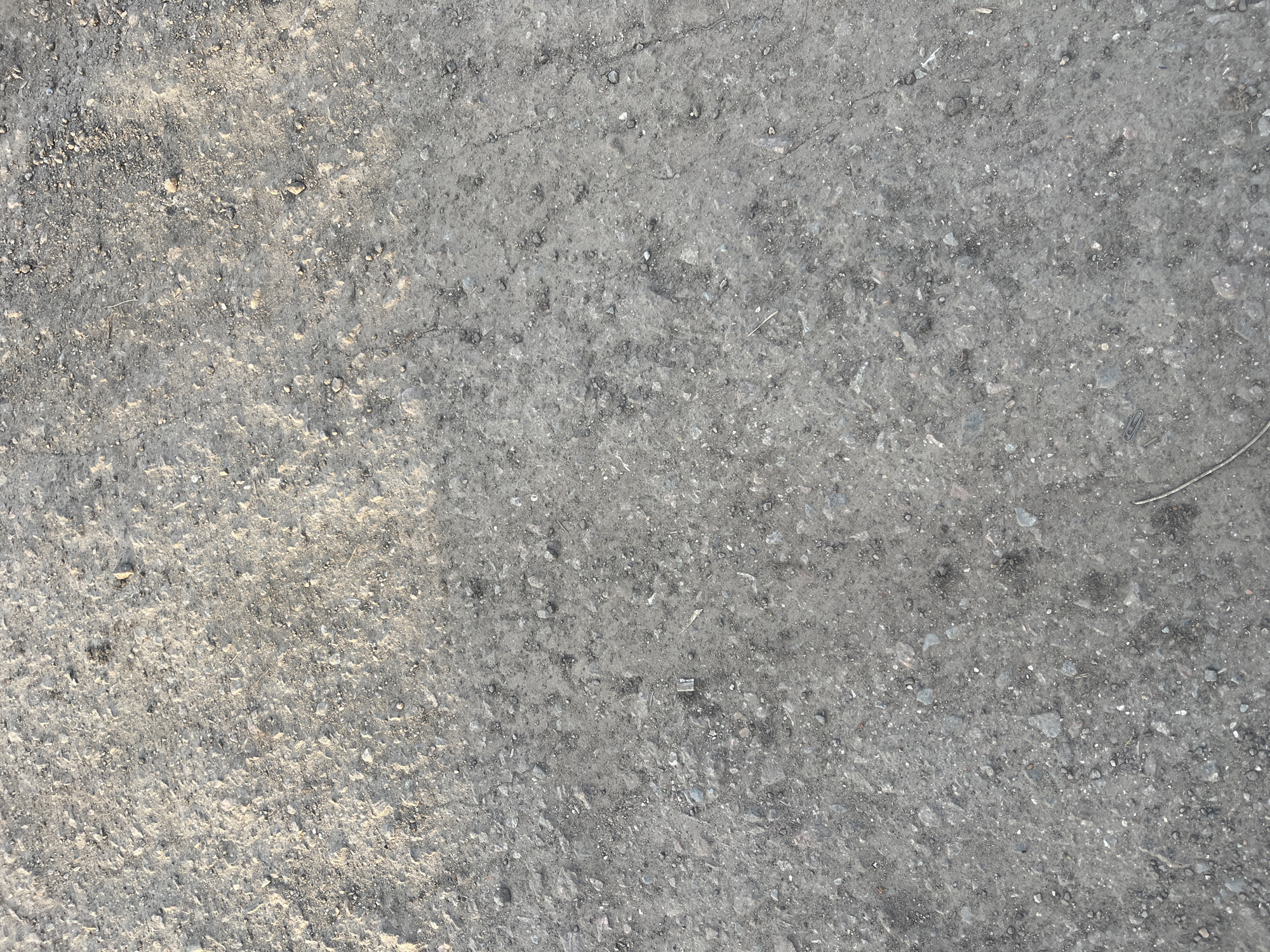 The image shows a close-up of a gray asphalt surface, likely a road or pavement. The surface is speckled with small stones and debris. The lighting is uneven, with brighter areas on the left side of the image. The image provides no information about the location being Podolsk, Russia, and there are no people, subjects, objects, actions, or interactions visible.