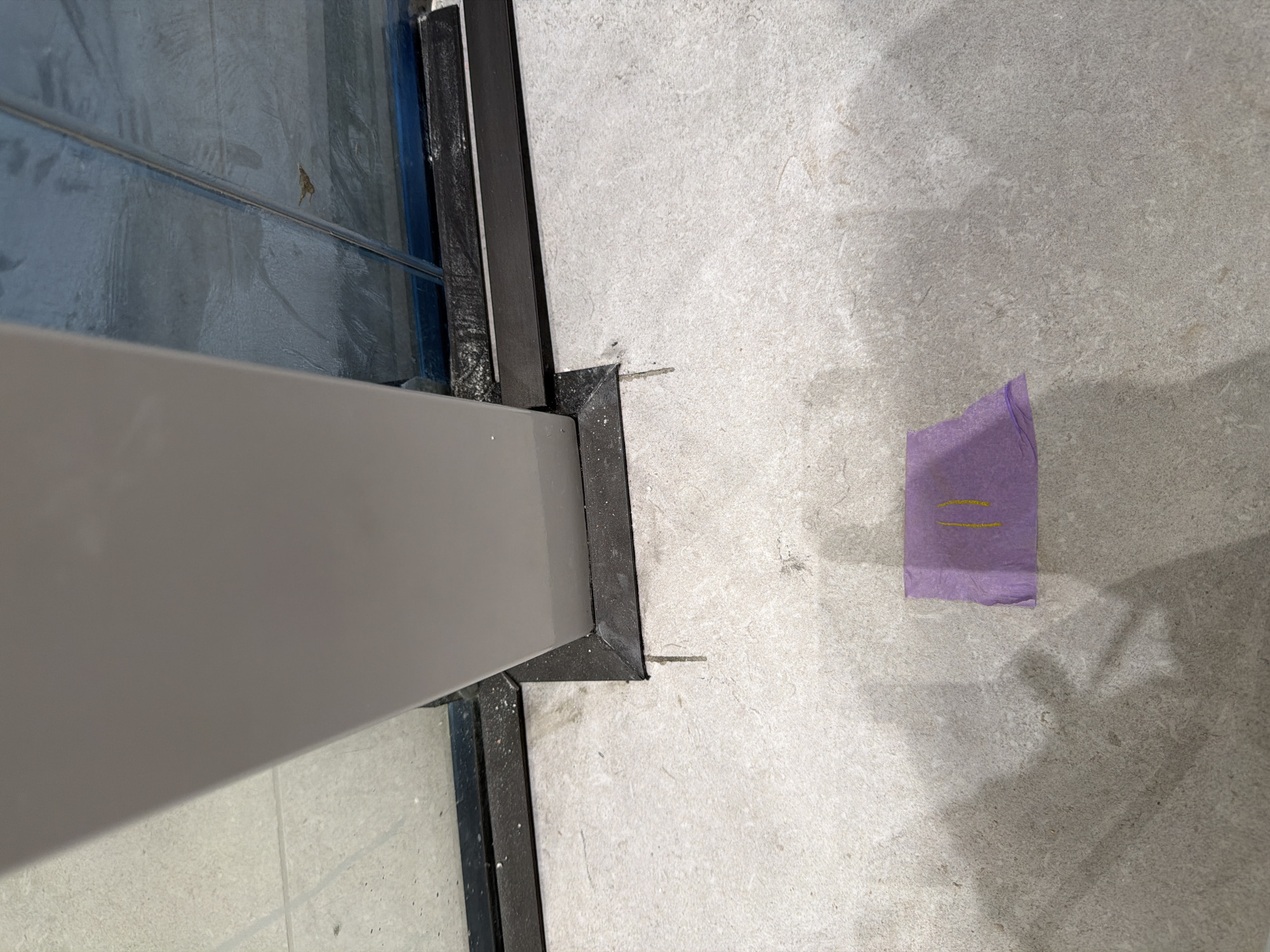 The image shows a close-up of a light grey tiled floor with a vertical, light grey rectangular post extending from the floor towards the top of the frame. The post is encased at its base by a dark grey material. To the right of the post, there is a section of glass, covered with blue protective film. The glass appears to be part of a larger structure, possibly a partition or window. On the floor, in the lower half of the image, is a small piece of purple tape with two yellow lines drawn on it, resembling the number "11." The scene is indoors, and the lighting suggests a well-lit environment, possibly commercial or residential. There are shadows cast on the floor, indicating a light source from the upper left. No people are visible in the image. The location is stated to be Jerusalem, Israel.