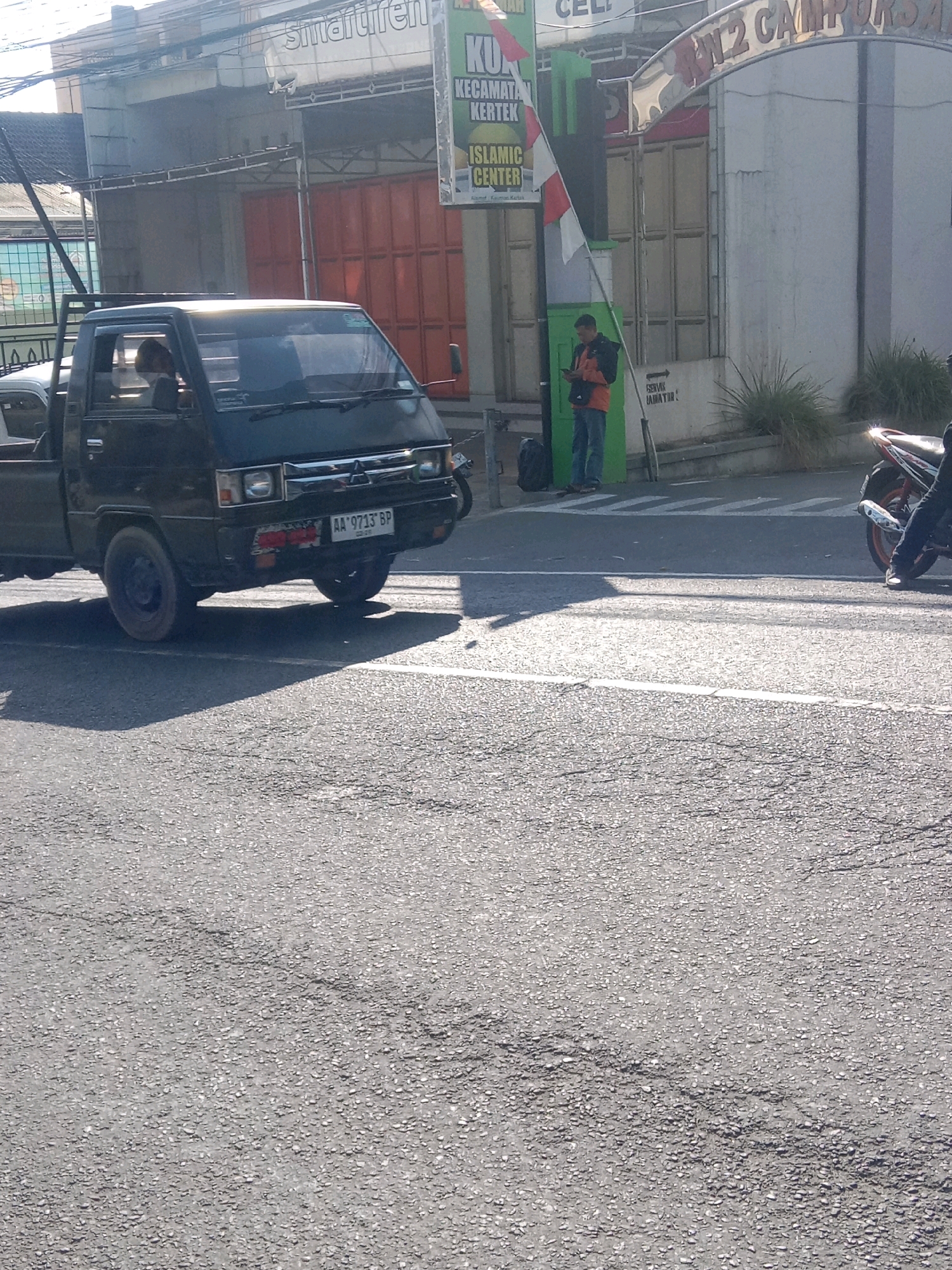 A black pickup truck with license plate "AA 9713 BP" is parked on a street in Kertek, Indonesia. The truck is facing left and a person is visible inside the driver's seat. In the background, a sign reads "KECAMATAN KERTEK" and "ISLAMIC CENTER". A person in an orange shirt and blue jeans is standing on the sidewalk next to a flag. A motorcycle is parked to the right. The street is paved with asphalt.