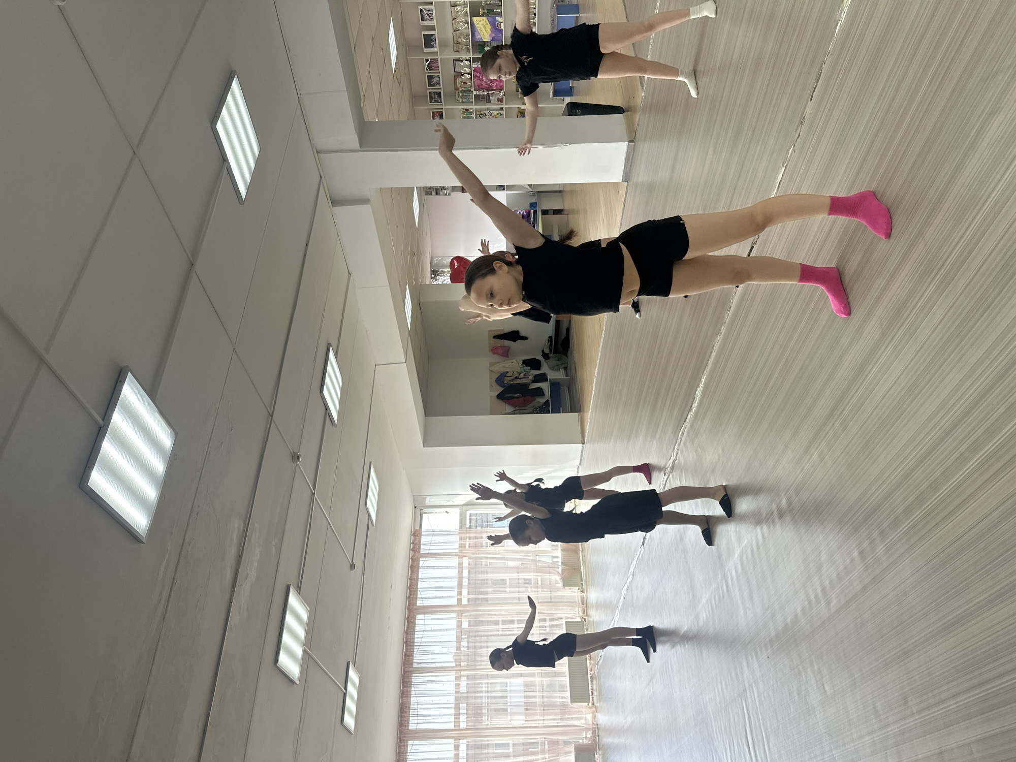 The photograph captures a dance class in progress. The dancers, dressed in black tops and shorts, are in various stages of a synchronized routine. One dancer in the center has pink socks and a red bow in her hair. The setting appears to be a dance studio with light-colored wooden floors and white walls lined with rectangular light fixtures.
Based on the information provided, the image was taken in the city of Votkinsk, Russia. Further specific location information isn't possible from the provided image alone.