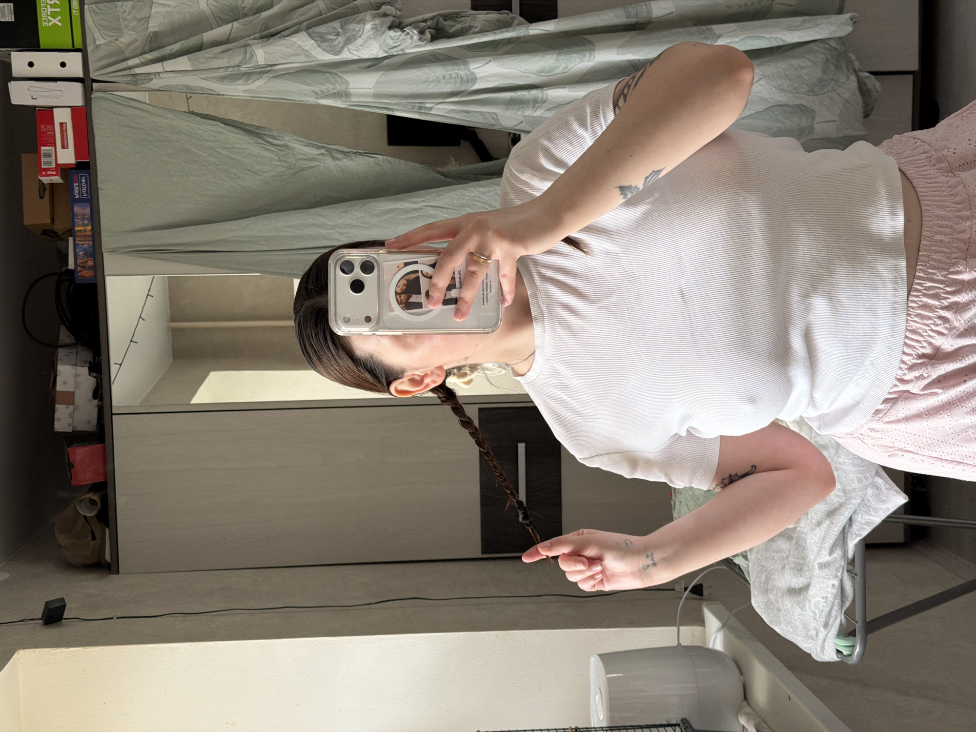 The image, taken in Chaikovsky, Russia, depicts a person capturing a mirror selfie indoors. The photograph is rotated 90 degrees counter-clockwise. The subject, a person with dark hair, is seen from the chest up, wearing a white ribbed short-sleeved top and pink, textured shorts. Their dark hair is styled into a single braid, secured with two black elastic bands, extending over their left shoulder and held by their left hand. A golden ring is visible on their right ring finger. Tattoos are present on their left upper arm (resembling a butterfly or bird) and a smaller, indistinct design on their left wrist.
The person holds an iPhone in their right hand, angled to reflect their image in a large mirror. The phone case is clear, revealing multiple circular images or photos on its back.
The background setting appears to be a domestic space. To the left, a vertical shelving unit holds various boxes, including a green box with "RTX" and "XTR" text, a red box labeled "Redmond," and a blue box marked "jigsaw." Light green curtains with a leaf pattern are visible behind the person. The lower portion of the scene includes a light-colored floor, a black power adapter with a cable, and a white appliance, possibly a fan or humidifier. The natural light suggests it is daytime.