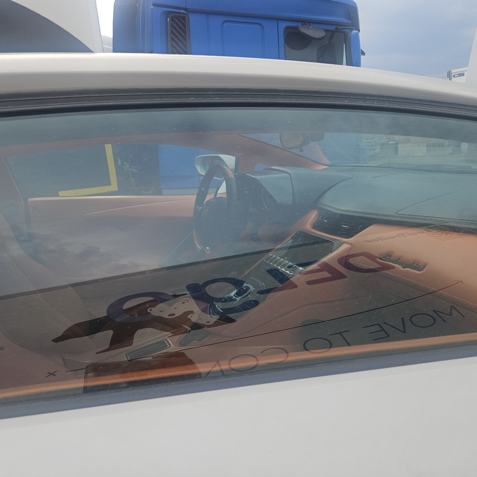 The image captures the interior of a luxury sports car, possibly a Lamborghini, seen through its windshield. The car's interior is upholstered in a light orange material. A steering wheel is visible, along with parts of the dashboard and the center console. A reflection on the windshield shows a blue truck and the sky. Text is superimposed on the image, with the words "DRAG" and "MOVE TO COM" visible, along with a small "Lamborghini" logo embossed on the dashboard. The car appears to be parked outdoors, as suggested by the truck in the reflection and the bright sky. The location is identified as La Jonquera, Spain.