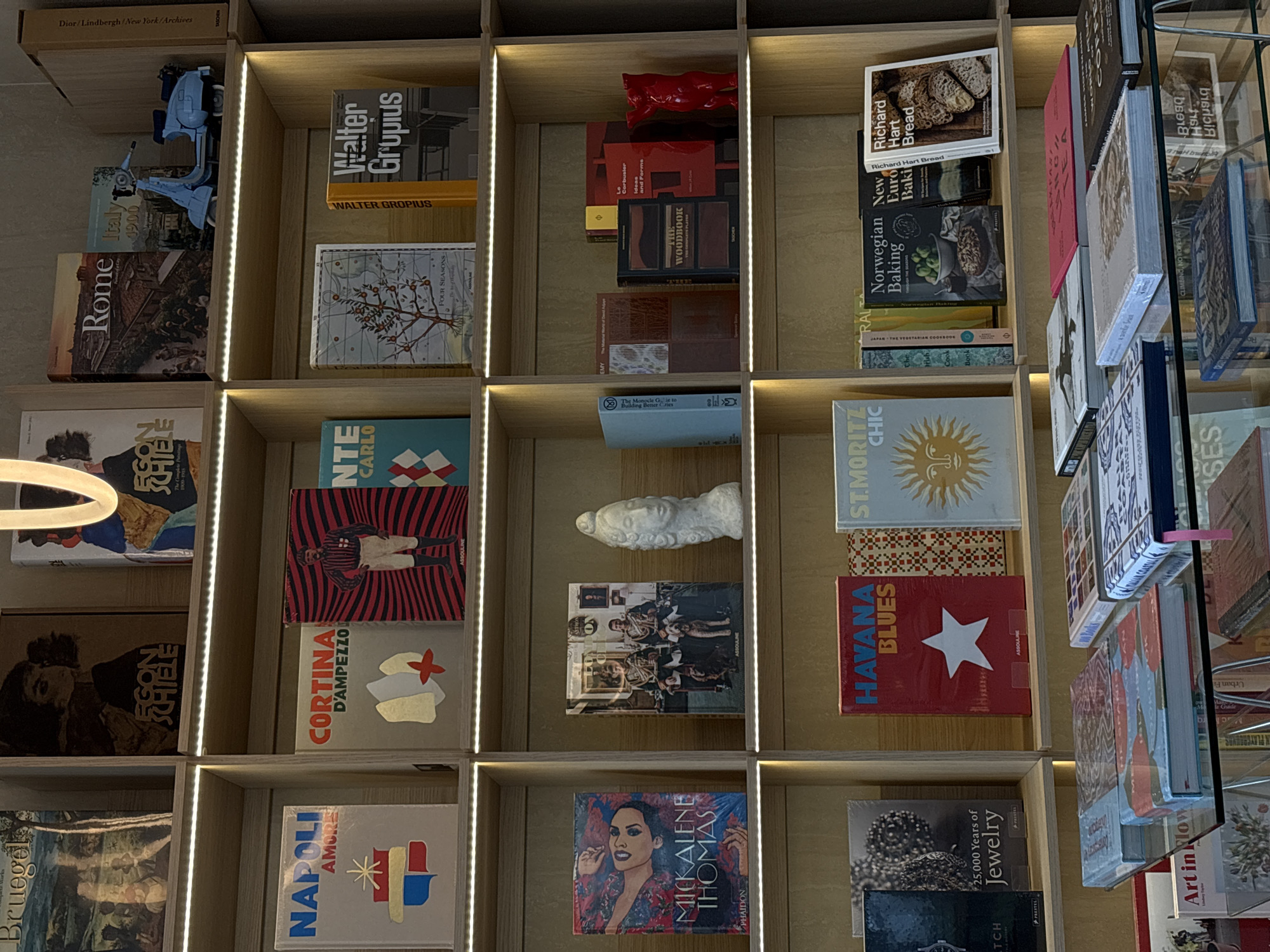 The image shows a well-lit bookshelf filled with various books and a few decorative objects. The books are arranged in individual compartments, creating a grid-like pattern. Titles visible include "Rome," "Italy 1900," "Egon Schiele," "Bruegel," "Napoli Amore," "Cortina D'Ampezzo," "Mickalene Thomas," "25,000 Years of Jewelry," "Havana Blues," "St. Moritz Chic," "Richard Hart Bread," "New Euro Baki," "Norwegian Baking," "Walter Gropius," "Four Seasons," and "The Woodbook."
Additional objects on the shelves include a red abstract sculpture, a white bust sculpture, a toy Vespa scooter, and other unidentifiable items. The lighting appears to be integrated into the bookshelf design. The shelves are clean and the overall presentation is curated and organized. There is no indication of Minsk, Belarus or any specific location in this image. There are no people visible in the image.