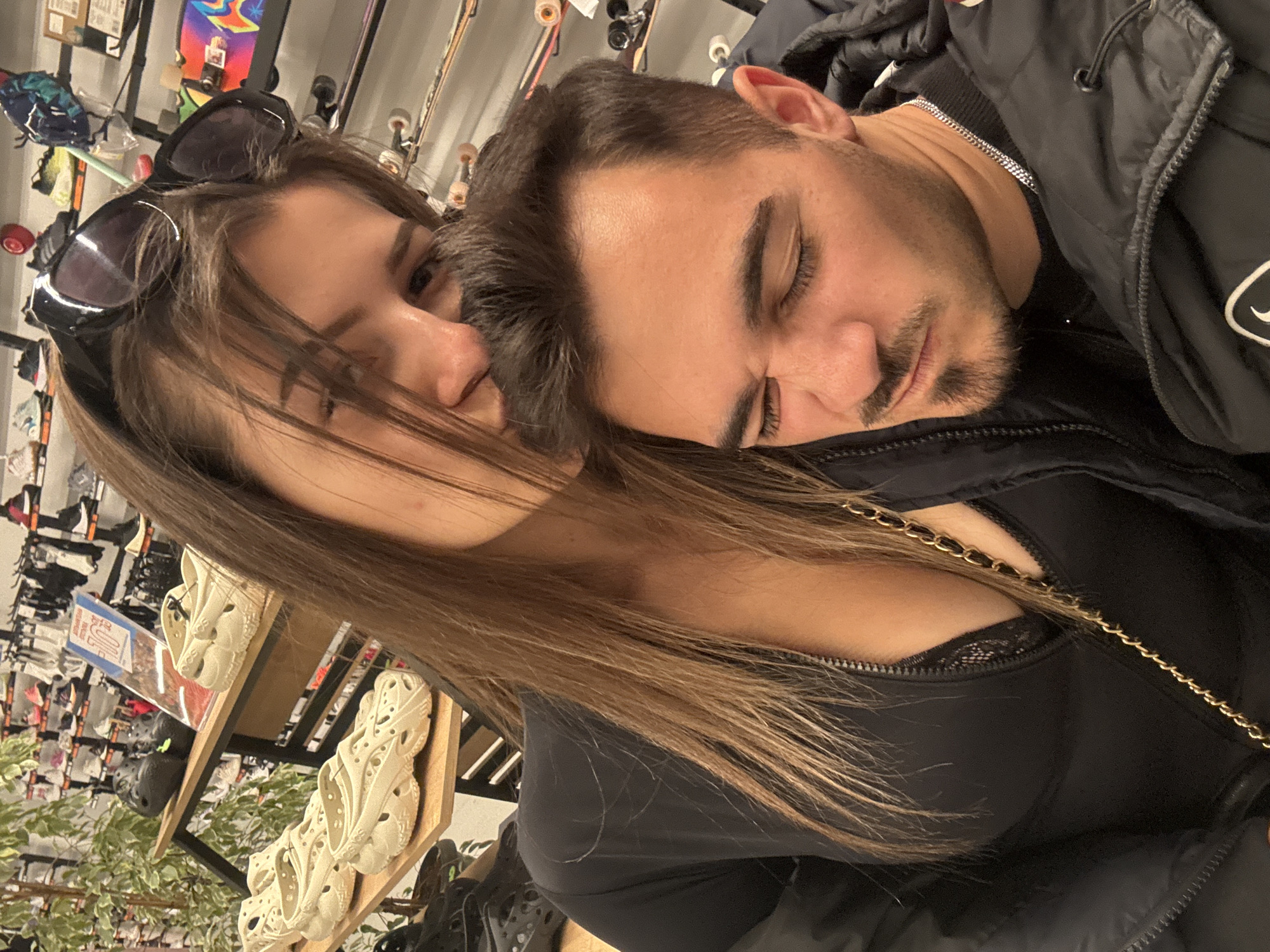 An overhead, slightly tilted shot captures two individuals, a man and a woman, in an indoor retail setting, likely a sporting goods or fashion store, given the context of Sofia, Bulgaria. The man, with short dark hair, a beard, and wearing a black puffer jacket over a silver chain necklace, is resting his head on the woman's shoulder/chest, with his eyes closed, suggesting sleep or profound relaxation. The woman, with long brown hair and sunglasses perched on her head, is looking directly at the camera with a neutral expression. She is wearing a black top, possibly a camisole with lace details visible, under a black jacket, and has a gold chain strap from a bag visible near her chest.
The background reveals rows of shelves stocked with various merchandise. Several pairs of light-colored, intricately designed clogs or sandals, reminiscent of Crocs, are prominently displayed on a wooden shelf next to a leafy green plant. Further back, skateboards with diverse graphics line the walls, and other sports equipment or apparel can be discerned. A blue sign in the background near the shoes partially displays "SUPER SALE," indicating a commercial environment. The overall mood conveys intimacy and comfort between the two subjects amidst a casual shopping backdrop.