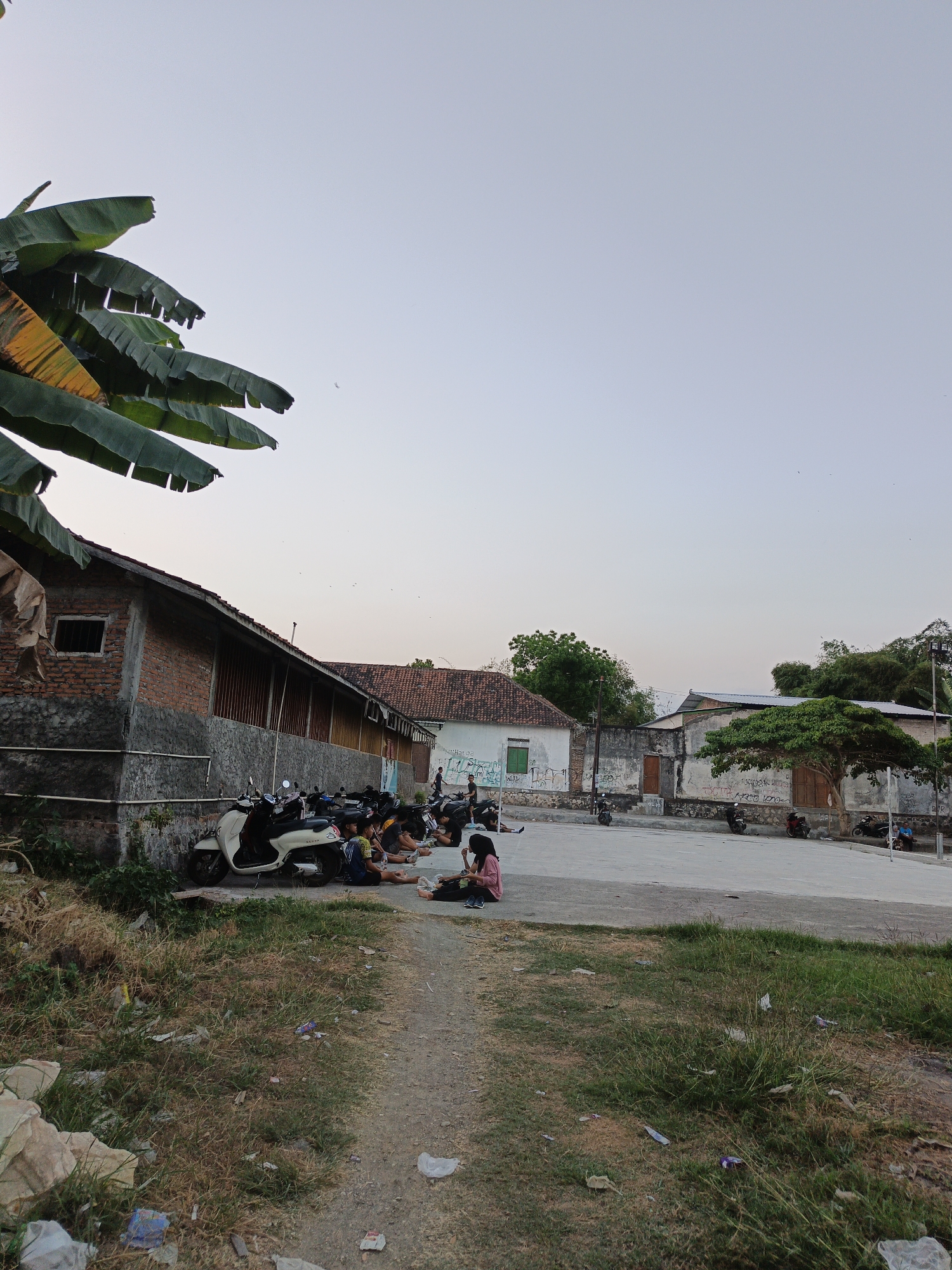 The image depicts an outdoor scene in Planggu, Indonesia, with a clear, pale sky dominating the upper portion. In the foreground, dry, overgrown grass and scattered debris line a dirt path leading into the frame. A red brick building with a stone base and a small, barred window stands on the left, with several motorcycles parked along its wall. Beyond the building, a paved area is visible, where a group of individuals, appearing to be mostly young people, are seated on the ground. Some are interacting with each other, while others are looking at their phones. Various motorcycles are parked in this area as well. Further in the background, more buildings with tiled roofs and a large tree with a sprawling canopy are present. The overall impression is a communal outdoor gathering space in a rural or semi-urban setting.