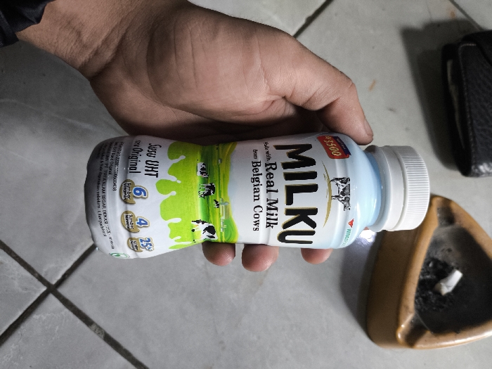 An adult's right hand holds a white plastic 'MILKU' brand UHT original milk bottle, featuring a white screw cap. The bottle label displays "Susu UHT Original," "Made with Real Milk from Belgian Cows," a cow graphic, green splatters, and a "Rp 3500" price tag. The scene is set on a light-colored tiled surface with dark grout lines. To the right of the bottle, a brown ceramic ashtray contains cigarette ash and a single discarded cigarette butt. Further right, a portion of a dark-colored wallet or small bag is visible. The location is Cibatu, Indonesia.