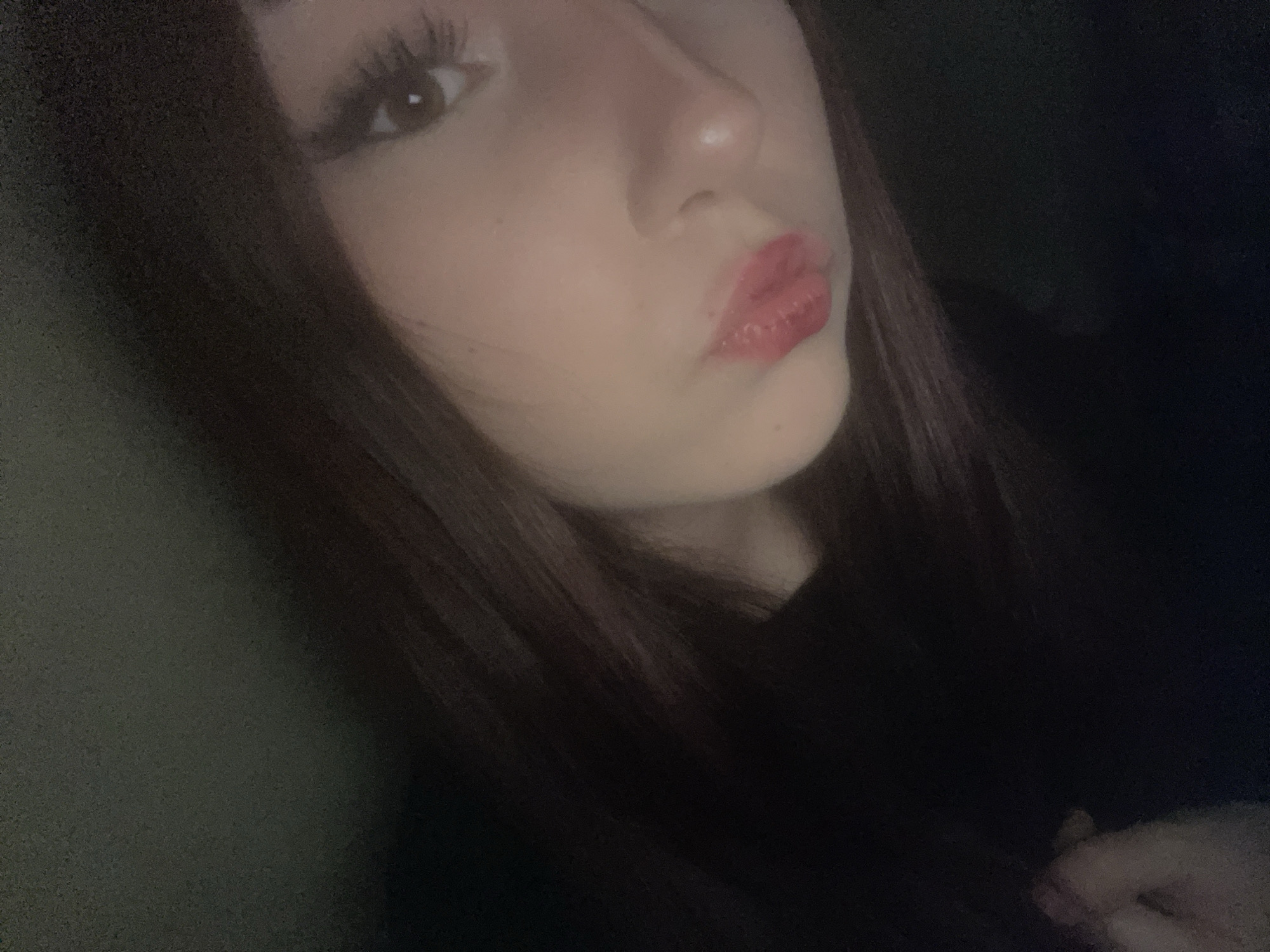A close-up, angled portrait features the right side of a young person's face against a dark, indistinct background. The subject, likely female, has long, dark hair that frames her face and extends out of view. Her right eye is visible, looking towards the camera, with prominent dark eyelashes, suggesting makeup. Her nose is partially seen, and her lips are puckered in a kissy face or pout, enhanced with glossy red or pink lipstick. The skin appears fair. The lighting is dim and soft, creating a slightly grainy texture throughout the image, characteristic of low-light photography. The background is uniformly dark, offering no discernible details or location cues. No objects other than the subject's features and hair are identifiable. The activity is posing for a portrait photograph. No text is visible. The image offers no visual evidence to confirm the location context of "Saint Petersburg, Russia."