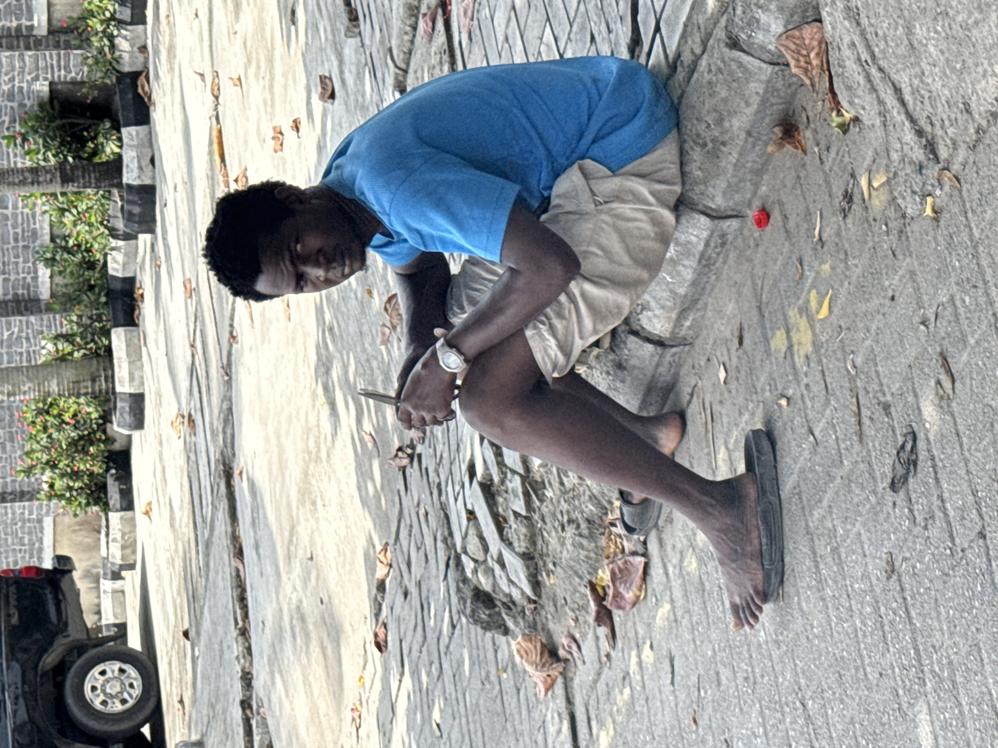A young man sits on a brick-paved walkway in Itirin, Nigeria. He wears a blue T-shirt and tan shorts. He holds a mobile phone in his hands. He is barefoot on one foot, while the other wears a black sandal. Debris litters the walkway, and a car is visible in the background.