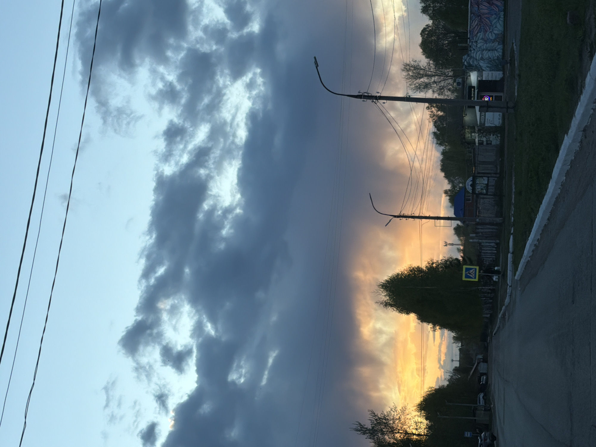 The image is a street scene taken in Chaikovsky, Russia, facing west towards a sunset. The sky is dominated by a bank of dark, gray storm clouds on the left, contrasting with a vibrant orange and yellow sunset on the right. Power lines stretch across the sky. Street lamps are visible along the road. A yellow pedestrian crossing sign is clearly visible on the right side of the road. A gray asphalt road occupies the foreground, with a partial view of the sidewalk on the right. Buildings, trees, and vegetation are visible along the road.