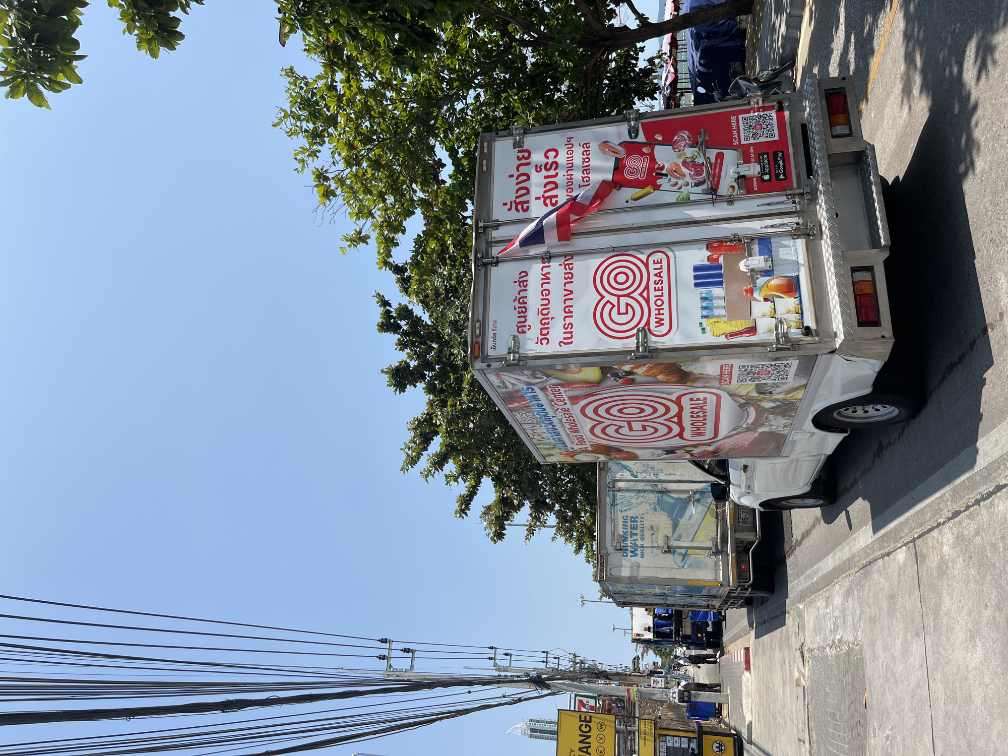 The image depicts a street scene in Pattaya City, Thailand. Several white commercial vehicles are parked along the roadside, their rear cargo areas adorned with advertisements for "GO Wholesale," a food wholesale center. The advertisements are in Thai and include QR codes for mobile app downloads. A small Thai flag is attached to one of the trucks. Overhead are numerous power lines crisscrossing the sky. A yellow sign with the text "CHANGE" is visible in the foreground. In the background, there are other vehicles and a few people. The sky is clear and blue, with tree branches visible above the trucks. The ground is a gray sidewalk and paved road.