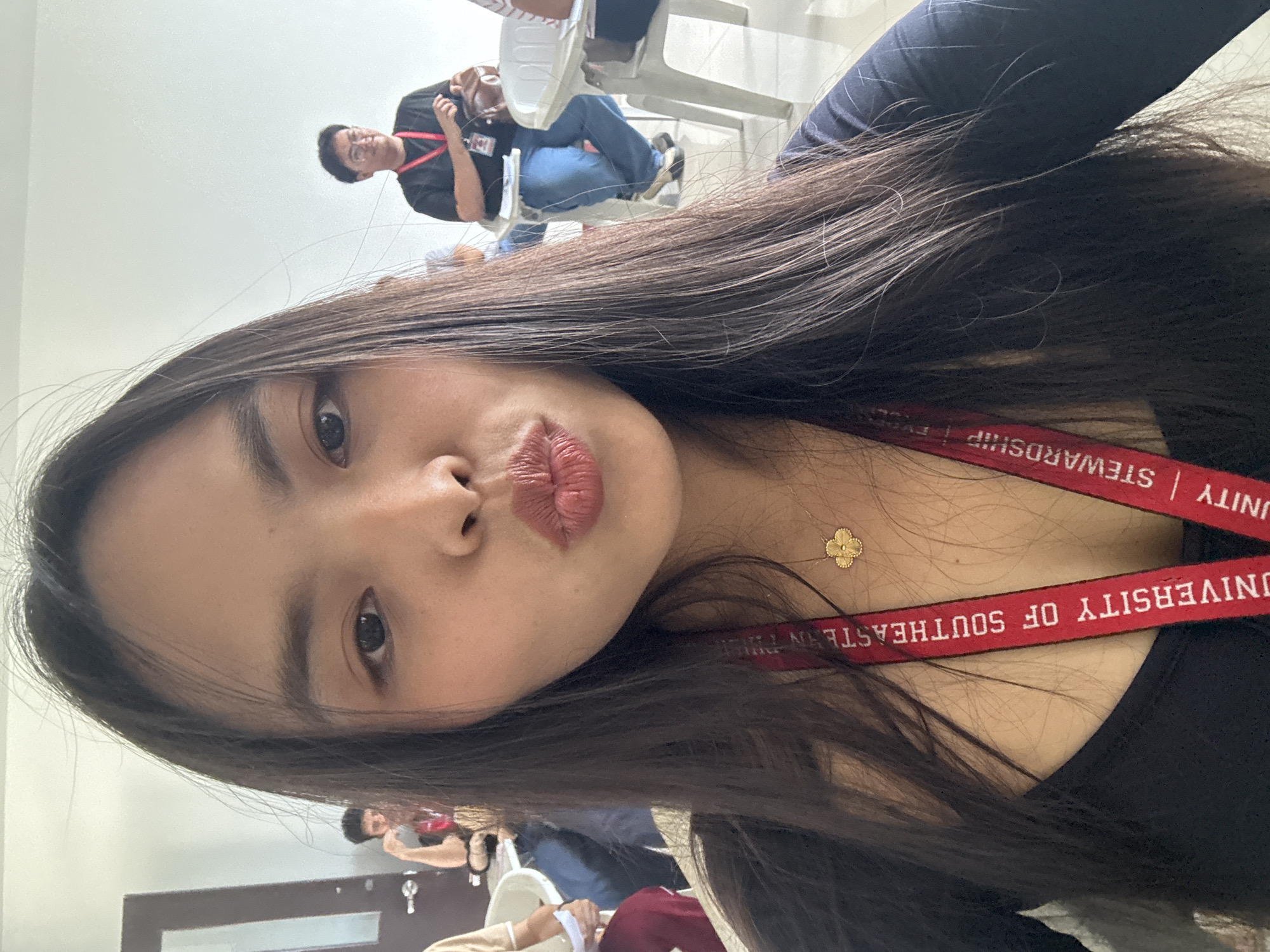 The image shows a selfie taken indoors by a young woman with long, dark hair. She is making a kissing face towards the camera. She is wearing a black top and a red lanyard with the words "UNIVERSITY OF SOUTHEASTERN PHILIPPINES | UNITY | STEWARDSHIP" visible. She also has a small gold flower necklace. In the background, other people are sitting on white plastic chairs. A man wearing a black shirt and jeans is seated in the background with a red lanyard. The location appears to be indoors, likely a classroom or conference room in Davao City, Philippines, indicated by the text on the lanyard. A door is visible in the back.
