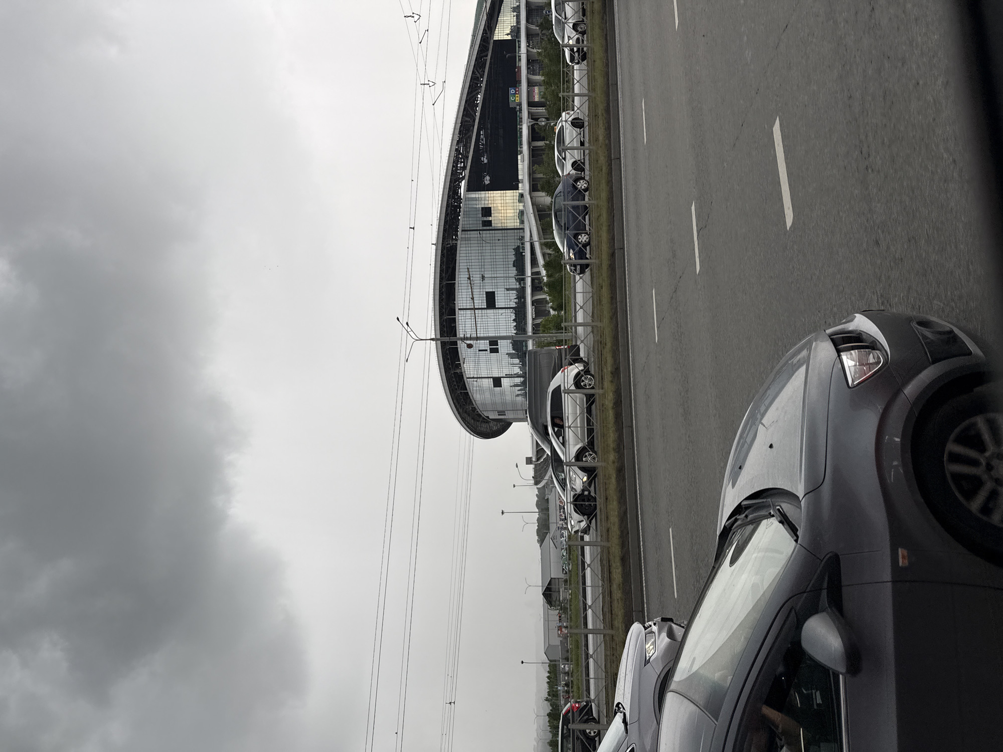 A grey, overcast sky looms over a traffic scene in Kazan, Russia. In the foreground, the hood and windshield of a dark grey car are visible, suggesting the photo was taken from inside another vehicle. Beyond this, a multi-lane road is congested with traffic. Several cars, including a white SUV, a dark blue sedan, and a white sedan, are stopped or moving slowly.
Further in the background, a modern stadium with a distinctive curved roof and a facade of reflective glass stands prominently. The stadium's architecture suggests a large public venue. Behind the stadium, the outline of other buildings and some trees can be seen against the hazy horizon. Power lines stretch across the upper portion of the frame, adding to the urban landscape. The overall impression is of a busy city environment under a gloomy sky.