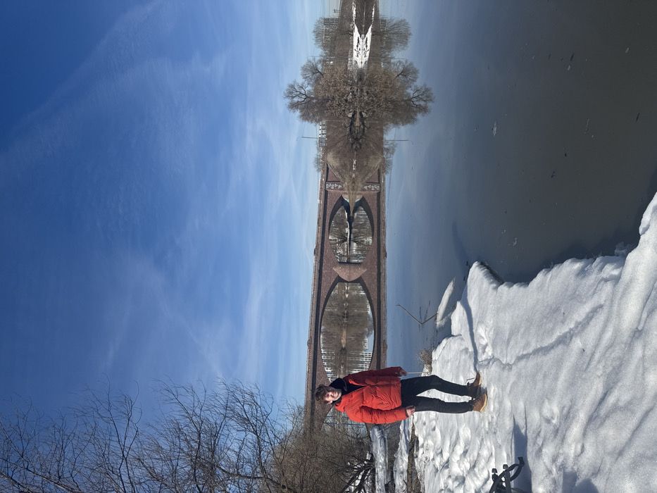 A wide-angle outdoor photograph captures a young, light-skinned male subject standing on a snow-covered riverbank under a bright blue sky. The subject, positioned on the left, wears a red puffer jacket, dark trousers, and brown lace-up boots, facing the camera with a faint smile. Patches of melting snow line the bank, which transitions into a calm, dark river that dominates the right and central foreground. The river’s surface reflects a large, multi-arched reddish-brown bridge in the mid-background. The bridge, constructed of brick or stone, spans the river and carries vehicular traffic, with several cars visible on its deck. Its architectural details and reflections are prominent. Beyond the bridge, the far bank shows bare trees and distant urban structures. The sky overhead is clear blue with sparse, wispy clouds. The scene depicts a late winter or early spring environment in the city of Ям, Russia.