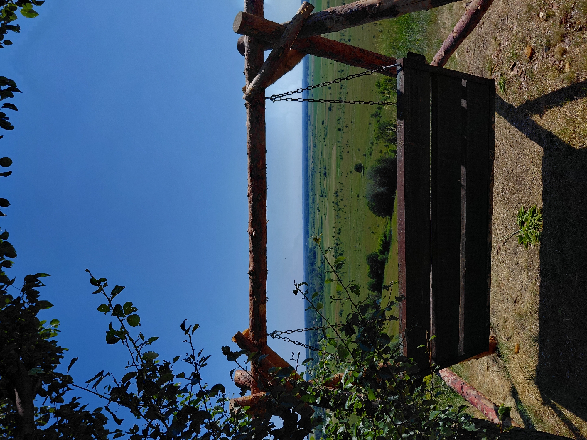 The image shows a wooden swing set with a dark wooden bench, suspended by chains from a wooden frame. The frame is constructed from rough-hewn logs. The swing is situated on a grassy area, with dry grass and a shadow cast by the swing frame indicating it is daytime, likely midday or afternoon given the strong shadow. Behind the swing, a vast, green, and somewhat undulating landscape stretches to the horizon, dotted with trees and fields. The sky above is a clear, deep blue. In the foreground, dark green foliage from a tree partially obscures the view. The scene suggests a rural or natural setting, possibly a park or a scenic viewpoint, with the Ukrainian countryside visible in the distance. There is no visible text in the image.