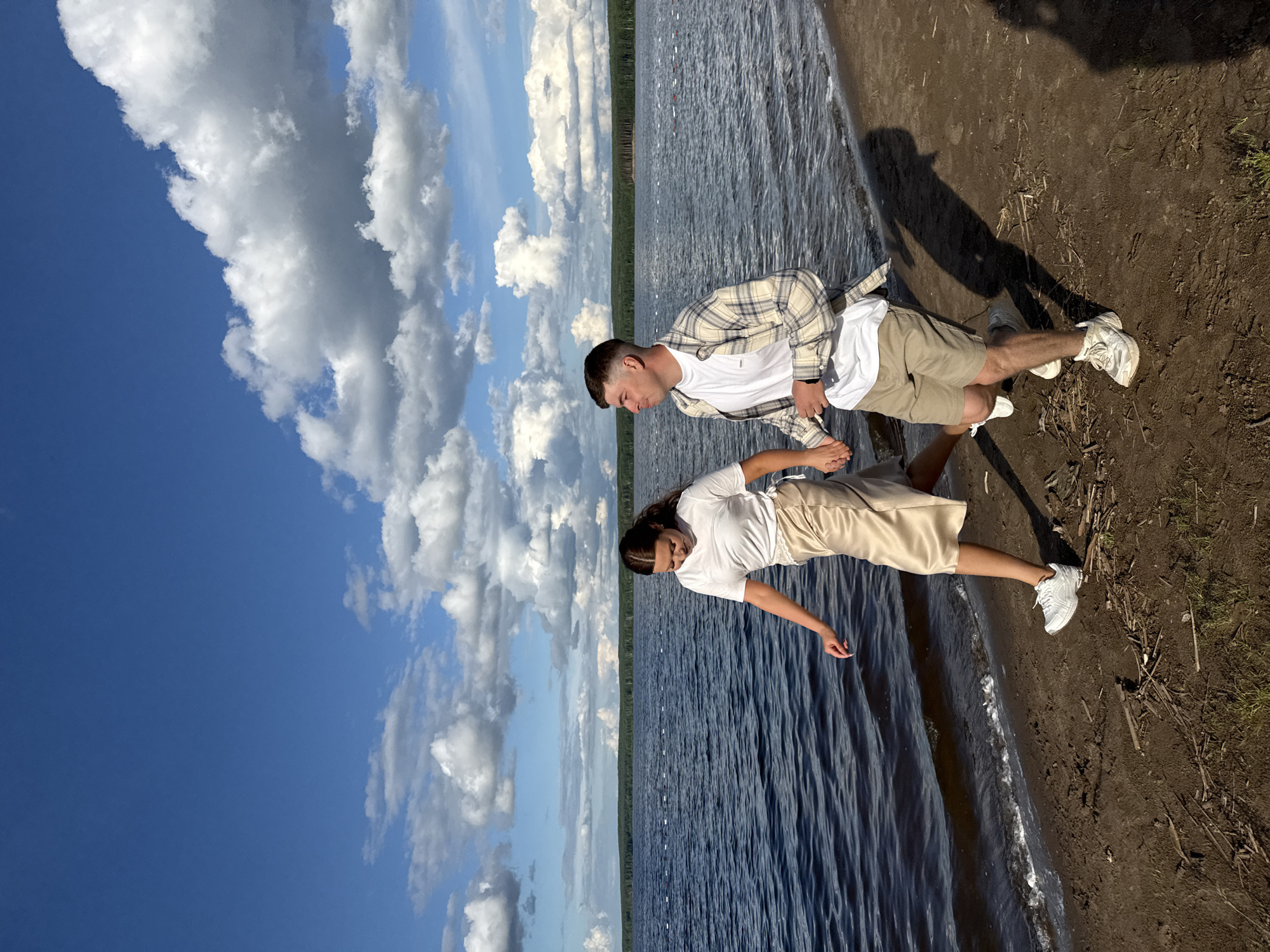 Two people, a man and a woman, stand on a shoreline near a body of water, presumably a reservoir or lake, under a sky with cumulus clouds. The man is wearing shorts, a white t-shirt, and a plaid overshirt. The woman is wearing a white t-shirt and knee-length shorts. Both are wearing white athletic shoes. The shoreline consists of dark dirt and water. In the distance, a line of trees is visible along the horizon. The location may be in or near Chaikovsky, Russia.