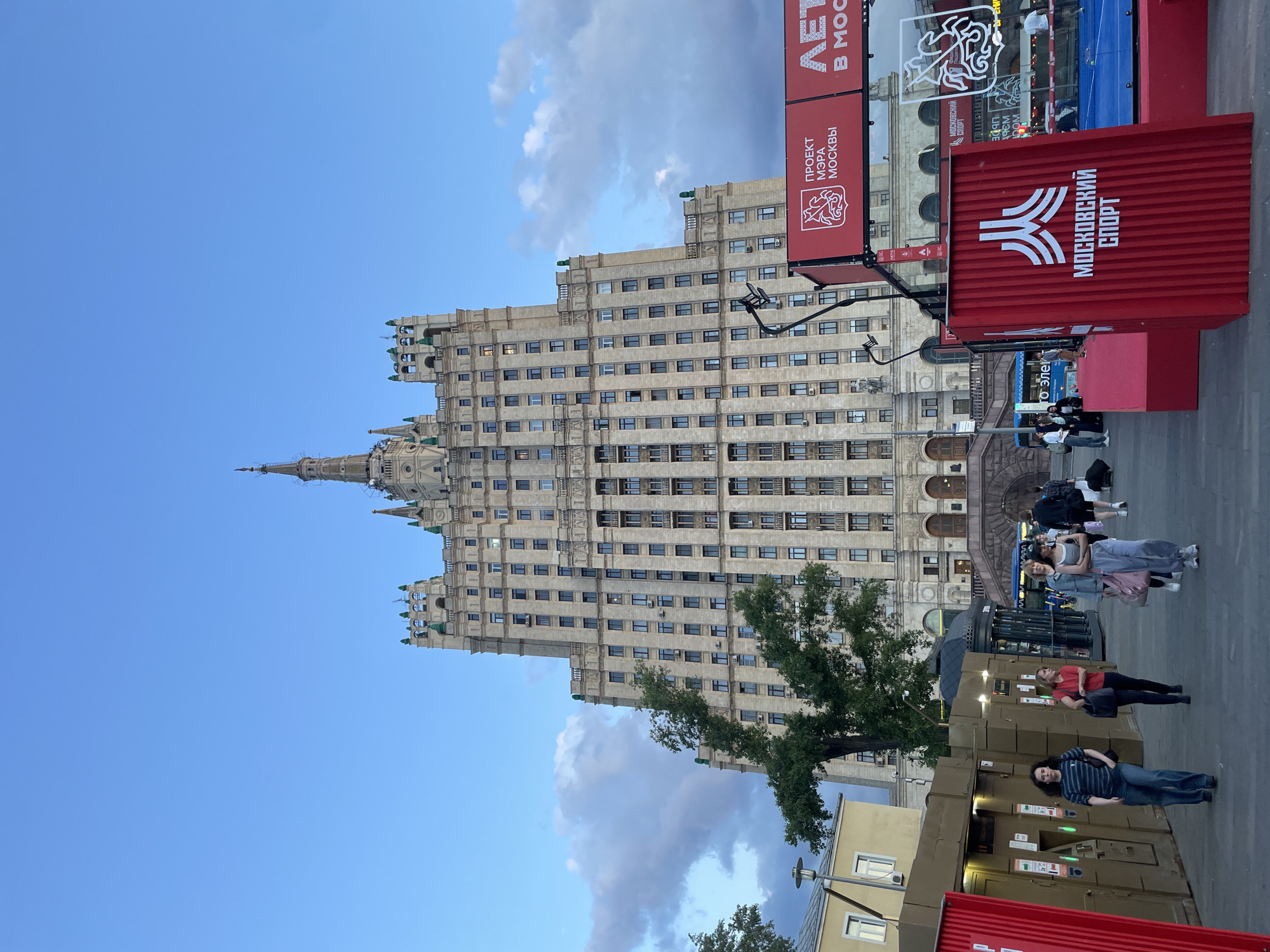 This media file captures a street scene in Moscow, Russia, dominated by a towering, ornate skyscraper, characteristic of Stalinist Empire architecture, similar to the "Seven Sisters." The image, initially rotated, depicts the building standing vertically against a clear blue sky with scattered white and grey clouds, suggesting late afternoon or early evening.
The main subject is a large, light-colored stone building with numerous rectangular windows spanning many stories. It features a prominent spire at its apex and several smaller decorative spires and green accents along its tiered roofline. A lush green tree partially obscures the lower-mid section of the building on the left.
At ground level, on the right side, there are several red banners and large, beige, box-like structures, possibly temporary kiosks or booths. Visible text in Cyrillic on the red banners reads "ПРОЕКТ МЭРА МОСКВЫ" (MOSCOW MAYOR'S PROJECT) and "МОСКОВСКИЙ СПОРТ" (MOSCOW SPORT), accompanied by a stylized logo. Another large red box features a white logo with three diagonal lines and the text "МОСКОВСКИЙ СПОРТ."
Several people are present at street level. One individual in a striped shirt and jeans leans against a box structure, while another in a red top and black pants stands nearby. Further back, a small group of people can be seen near what appears to be an entrance or steps to the main building. The overall scene conveys a typical urban setting with ongoing public activity, possibly related to a local sports initiative.