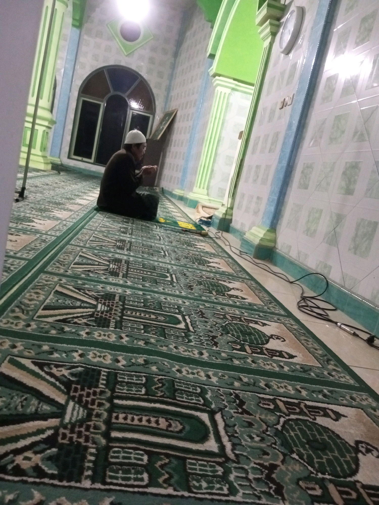 The image shows a man wearing a white cap and a dark robe, kneeling on a green and beige patterned carpet inside a mosque in Andongsili, Indonesia. The man is facing away from the camera and appears to be praying. He has his hands clasped together in front of him. The mosque has green pillars and tiled walls with a light blue trim. A clock is visible on the wall. The carpet has intricate geometric patterns and is laid out in rows. There are some cables on the floor near the wall. The lighting is somewhat dim, with a bright light source visible in the upper left corner of the image.