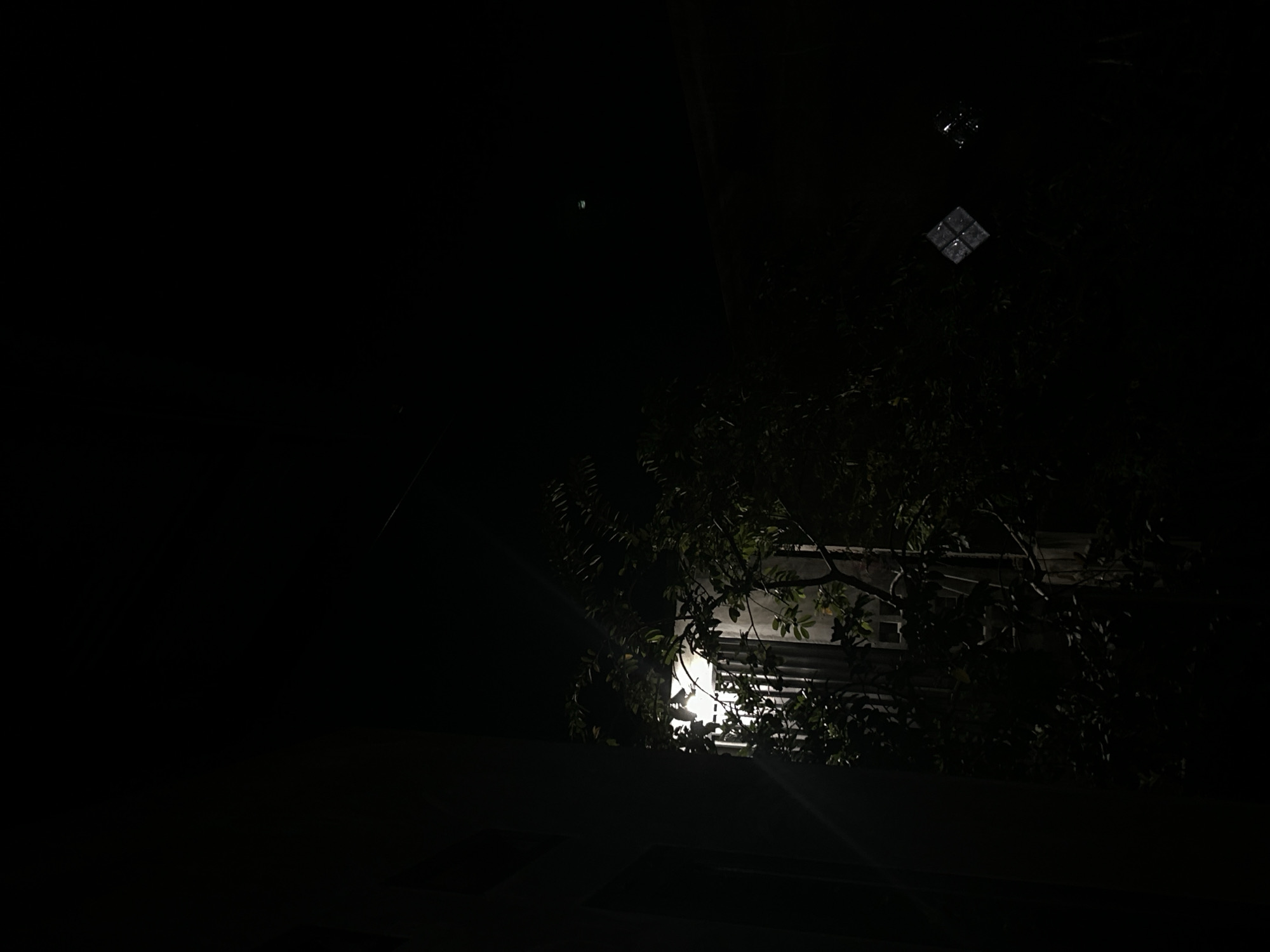 The image, taken at night, reveals a dark scene in Lhokseumawe, Indonesia. A building or structure is partially visible, illuminated by a bright light source that appears to be behind or within it, partially obscured by foliage. Above the building, there's a small, indistinct reflection, seemingly from a window. The light casts a flare, and the dark environment is mostly out of focus.