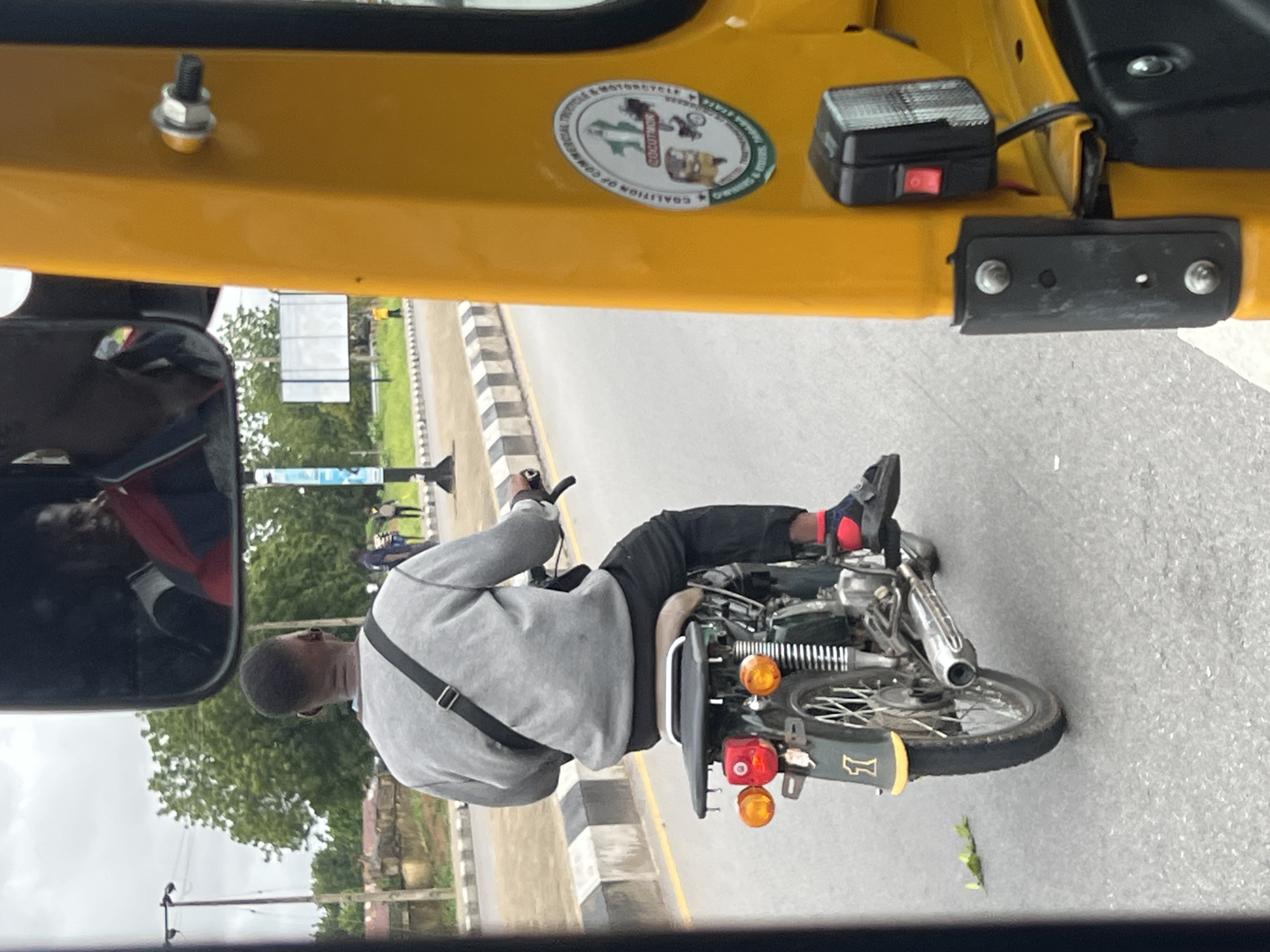 This image shows a close-up, side view of a man riding a motorcycle. The man is wearing a grey sweatshirt, black pants, and black shoes with red accents. He has a black strap across his chest. The motorcycle is dark green and appears to be older model. It has chrome accents and a visible exhaust pipe. The motorcycle has two orange turn signals and a red taillight. The man's feet are resting on the footpegs.
The scene is set on a paved road with a striped curb. In the background, there are trees and some buildings. There is a yellow structure, possibly part of a vehicle, in the foreground that frames the image. A reflection of another person wearing a red and blue jacket is visible in a mirror on the left side of the frame. A sticker is visible on the yellow structure, with text that appears to read "COALITION OF COMMERCIAL MOTORCYCLE". There is also a small black box with a red switch attached to the yellow structure.
The image appears to be taken during the day, with overcast or cloudy skies. The lighting is diffused, suggesting it might be a slightly dull day. The location is identified as Jalingo, Nigeria. There are no strong emotional cues evident from the man's posture or expression. The overall impression is a candid shot of someone commuting or traveling on a motorcycle in an urban or semi-urban environment in Nigeria.