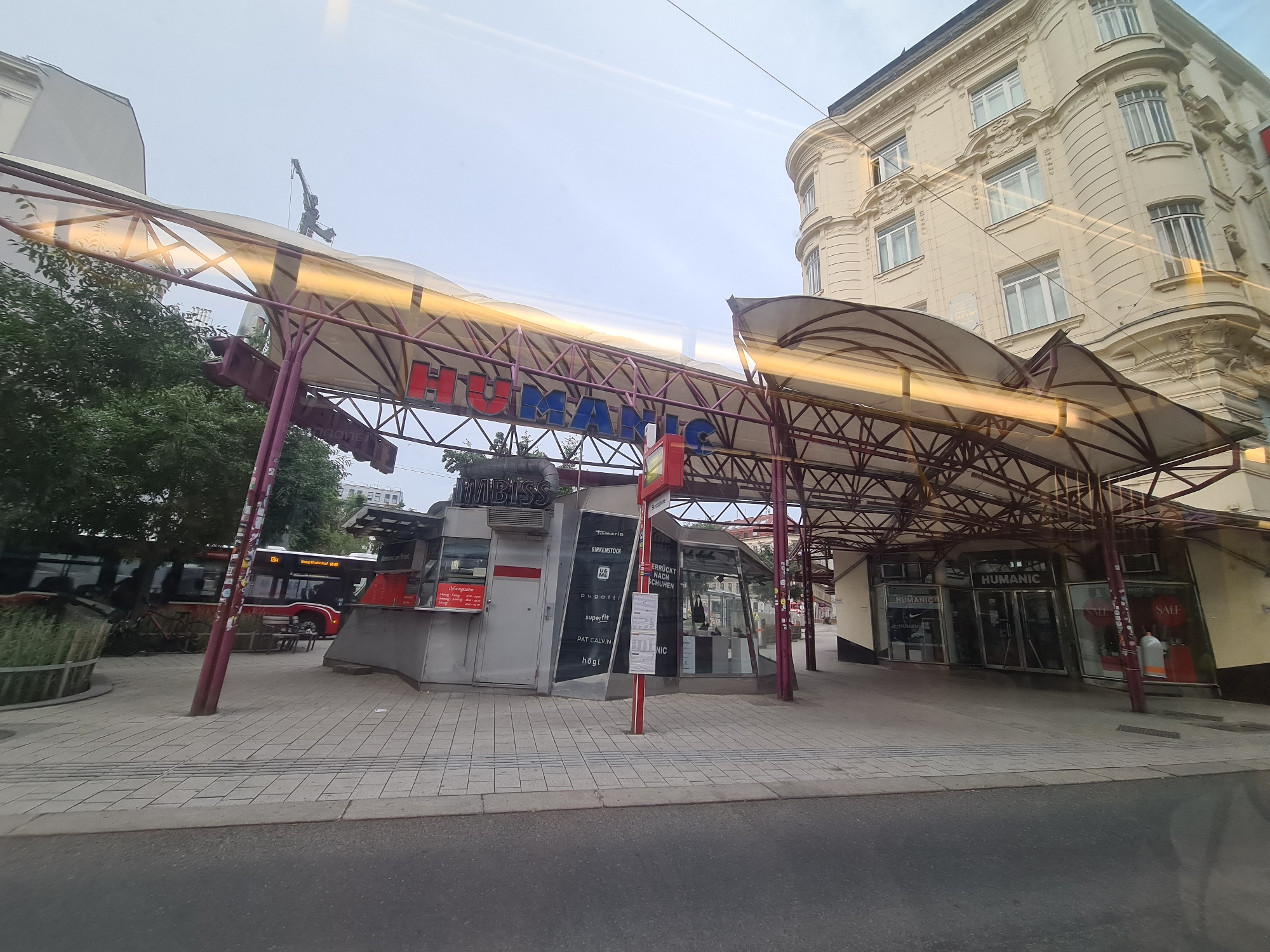 The image captures a street-level view of the "Am Graben" area in Vienna, Austria, specifically near the intersection with Annagasse. The foreground features a paved sidewalk with a visible crosswalk and a street. Overhead, a canopy structure with a purple frame and a translucent roof is present, extending over the sidewalk and partially obscuring the sky. Red neon lettering on the canopy spells out "HUMANIC".
To the left, a bus with the number "13A" prominently displayed is parked, its doors open, suggesting a bus stop. Adjacent to the bus, a kiosk with "IMBISS" (German for snack bar) signage is visible, displaying various brand names on its side, including Tamaris, Birkenstock, Bugatti, and Högl. Further to the right, a storefront with "HUMANIC" signage and large glass windows is visible, showcasing shoes and clothing. A "SALE" sign is also visible in the window.
The background includes a multi-story building with a light-colored facade, characterized by arched windows and decorative architectural elements. The sky is overcast, and a construction crane is visible in the distance, suggesting ongoing development in the city. No individuals are clearly visible or interacting within the frame. The scene appears to be a public thoroughfare with commercial establishments and public transportation.