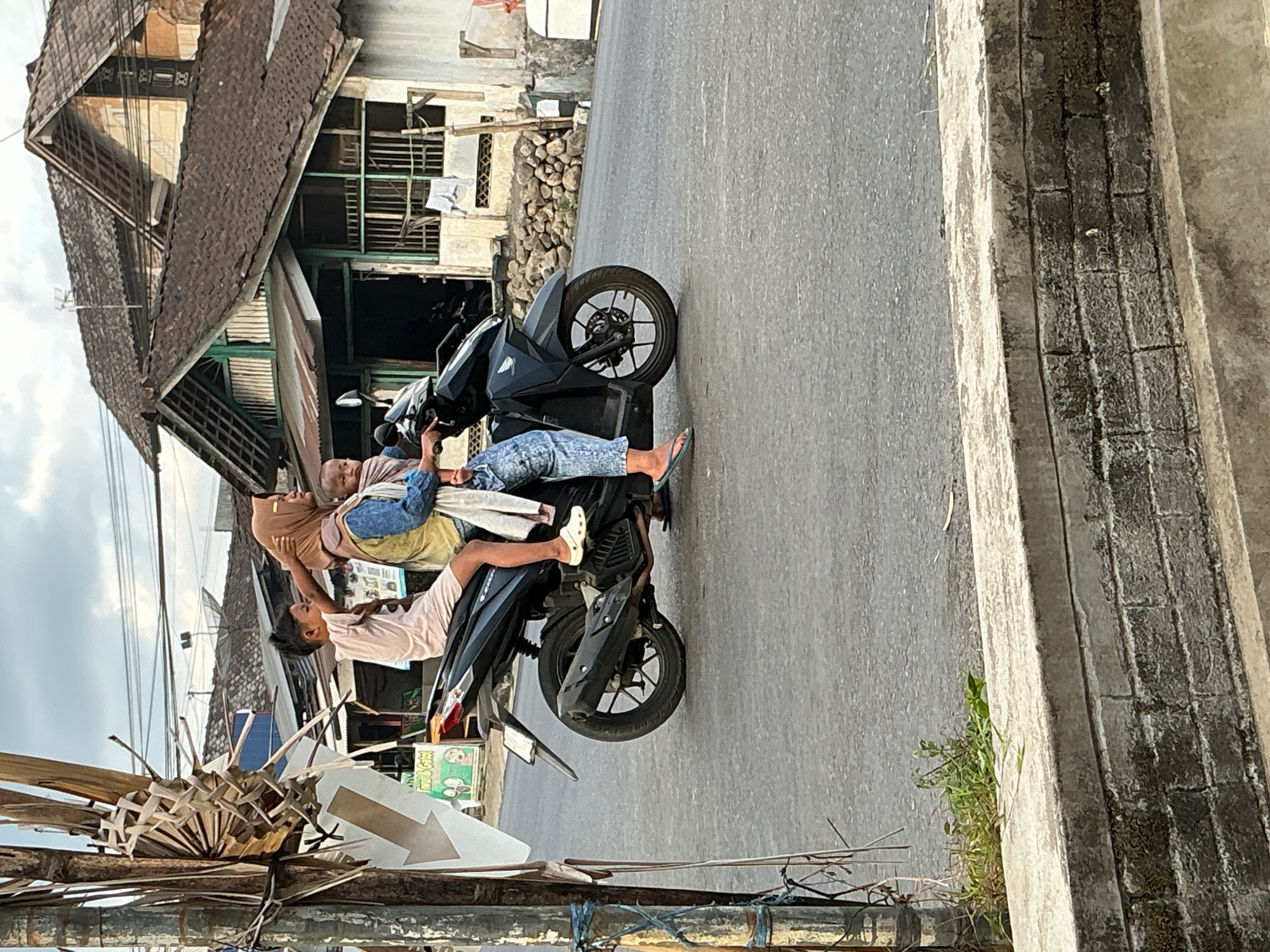 In Kebonrejo, Indonesia, a woman wearing a brown hijab and a blue jacket, along with a baby wrapped in a pink sling, rides on the back of a dark-colored motorcycle. A young boy, dressed in a light pink t-shirt and shorts, sits in front of her, appearing to adjust her hair. The motorcycle is moving on a paved road. In the background, there are several houses with brown tiled roofs and weathered walls. A tall pole with some woven material is visible on the left side of the image, near a traffic sign. The sky is overcast. A concrete barrier with visible brickwork is in the foreground.
