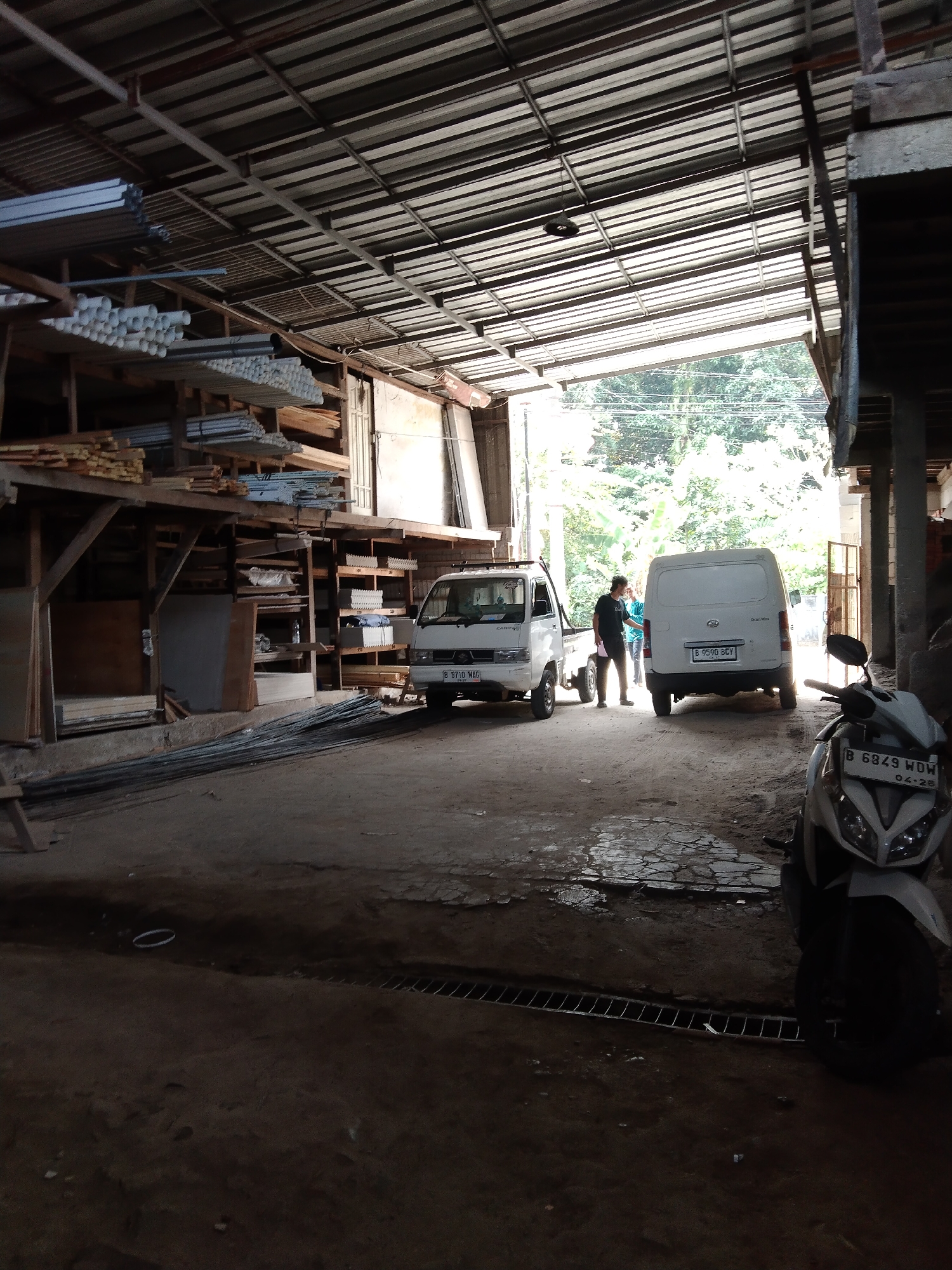 The image depicts an open-air warehouse setting, likely in Parung, Indonesia, characterized by its industrial and utilitarian nature. The primary structure features a corrugated metal roof supported by exposed beams and columns, allowing ample natural light to filter in from the open sides. Stacked materials, including lengths of metal pipes and wooden planks, are organized on shelves and on the floor, indicating a place for storage and distribution of construction or hardware supplies.
In the foreground, a parked white scooter with a license plate partially visible is positioned on a rough, cracked concrete surface. Behind it, two white vehicles are present: a small pickup truck and a panel van, suggesting recent activity or staging for transport. A man, dressed in a dark t-shirt and light-colored pants, stands between the two vehicles, possibly engaged in a conversation or transaction with another individual wearing a teal shirt who is facing away from the camera.
The environment suggests a functional, working space rather than a public area. The presence of construction materials, industrial vehicles, and the open-air layout are consistent with a warehousing or supply yard. The background shows a glimpse of greenery and trees, indicating the warehouse is situated in an area with surrounding vegetation. The overall lighting suggests it is daytime, with shadows cast across the ground, highlighting the uneven texture of the surface.
