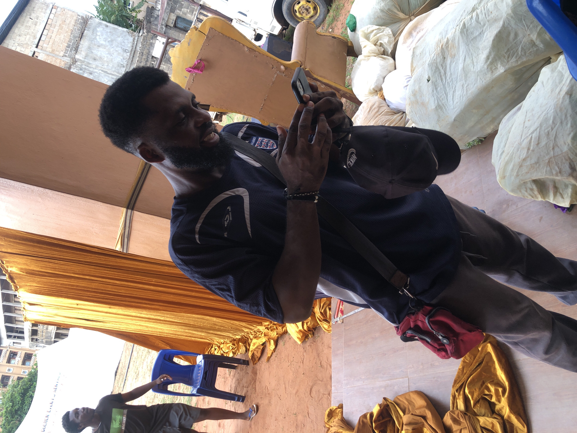 This image captures a vibrant, outdoor or semi-outdoor scene, likely in Onitsha, Nigeria, given the context.
The main subject is a dark-skinned man, positioned centrally, with a neatly trimmed beard and short dark hair. He is wearing a dark blue short-sleeved sports top with a white collar and a crest that reads "ENGLAND" above it, accompanied by white/silver design elements. He wears dark trousers and a black shoulder bag with a white "NIKE" logo, along with a small red fanny pack or pouch hanging from his side. A dark, beaded bracelet adorns his left wrist. He is actively engaged with a black smartphone, holding it with both hands and looking down at its screen. His expression suggests concentration or a slight smile.
In the lower left background, another dark-skinned man is visible, wearing a dark t-shirt with a green logo (resembling Lacoste) and dark shorts, paired with blue flip-flops. He stands casually next to a blue plastic chair.
The setting appears to be an event space, possibly under a large canopy or tent. Golden/yellow fabric drapes across the top of the frame and is also present on the ground. In the background, stacks of large, off-white or beige sacks are piled up. A dilapidated yellow wooden structure, resembling part of a counter or a prop, is positioned behind the main subject, with a vehicle wheel partially visible behind it. The ground transitions from exposed red dirt to a tiled or paved area. Further in the background, residential buildings, some appearing weathered or under construction, are visible, interspersed with greenery. A white banner or tent wall in the lower left background partially reveals the text "GOLDEN EVENTS RENTALS," confirming the event context.
The scene is lit by natural daylight, suggesting a daytime event. The atmosphere seems relaxed, with individuals either preparing for or taking a break during the event.