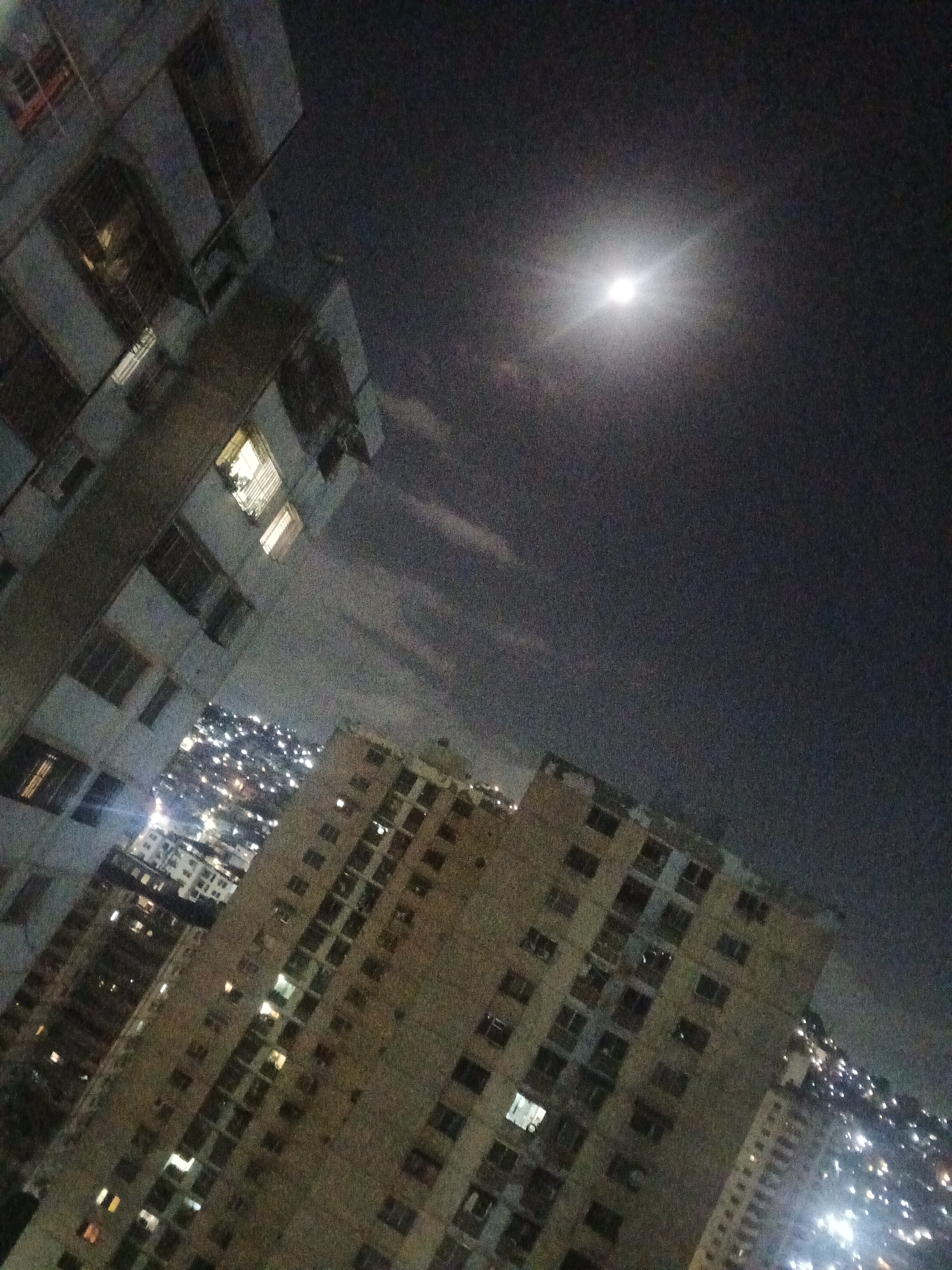 The image presents a nighttime view of Caracas, Venezuela, dominated by high-rise buildings under a moonlit sky. The buildings are illuminated by scattered lights, indicating occupancy. The sky is dark, with the moon as the primary light source, creating a contrasting effect. The buildings in the foreground appear to be residential apartments, constructed with a simple design. The overall atmosphere suggests a densely populated urban environment during nighttime hours. The image doesn't have any explicit people, subjects or actions.