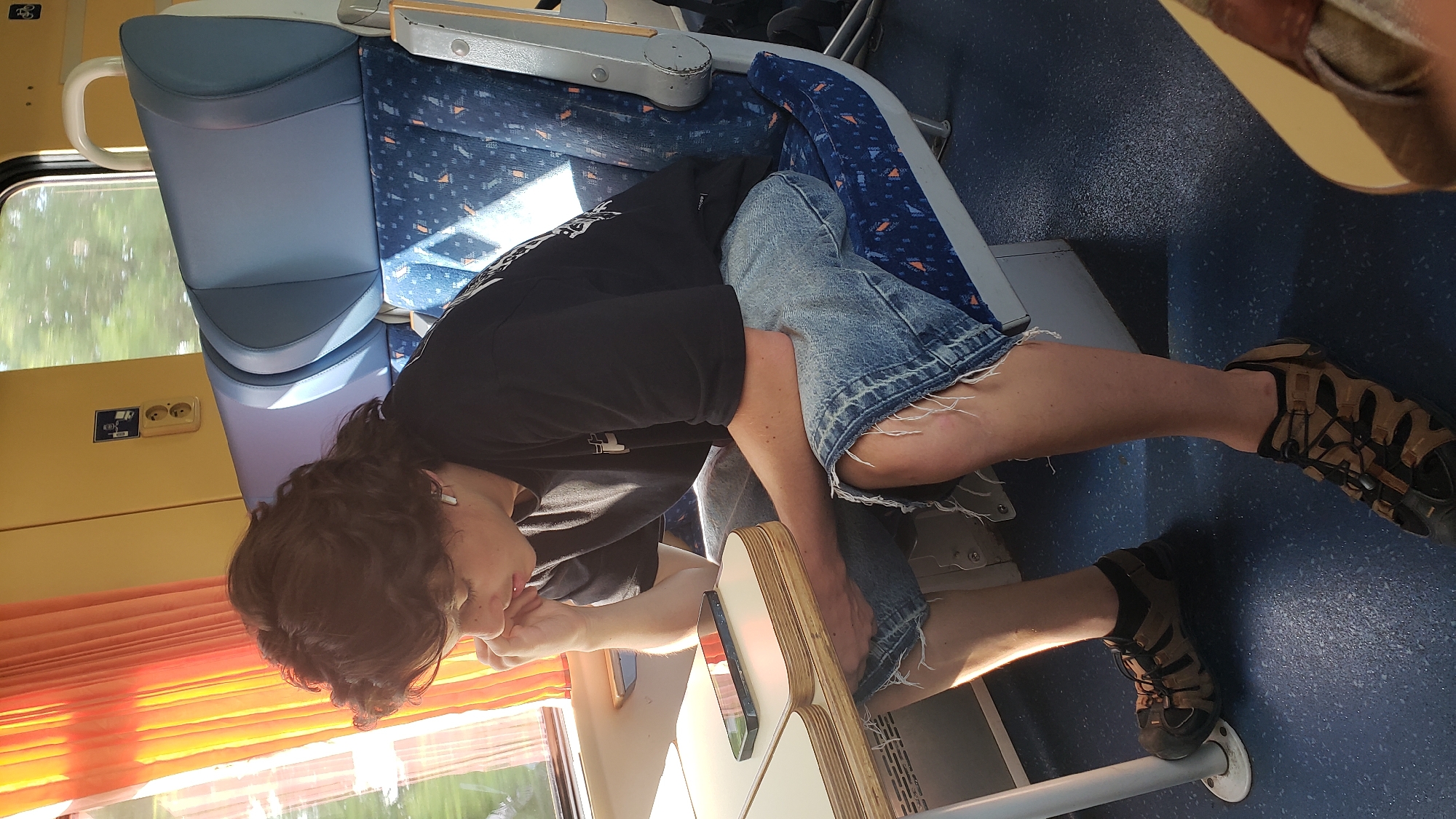 A person sits on a blue patterned seat in a train carriage, looking down at a mobile phone on a small table. The person has medium-length brown hair and is wearing a black t-shirt and distressed denim shorts. They are also wearing brown and black hiking sandals. The train carriage has orange curtains, yellow walls, and blue seats with a speckled pattern. Sunlight streams through the window, casting shadows on the seats.