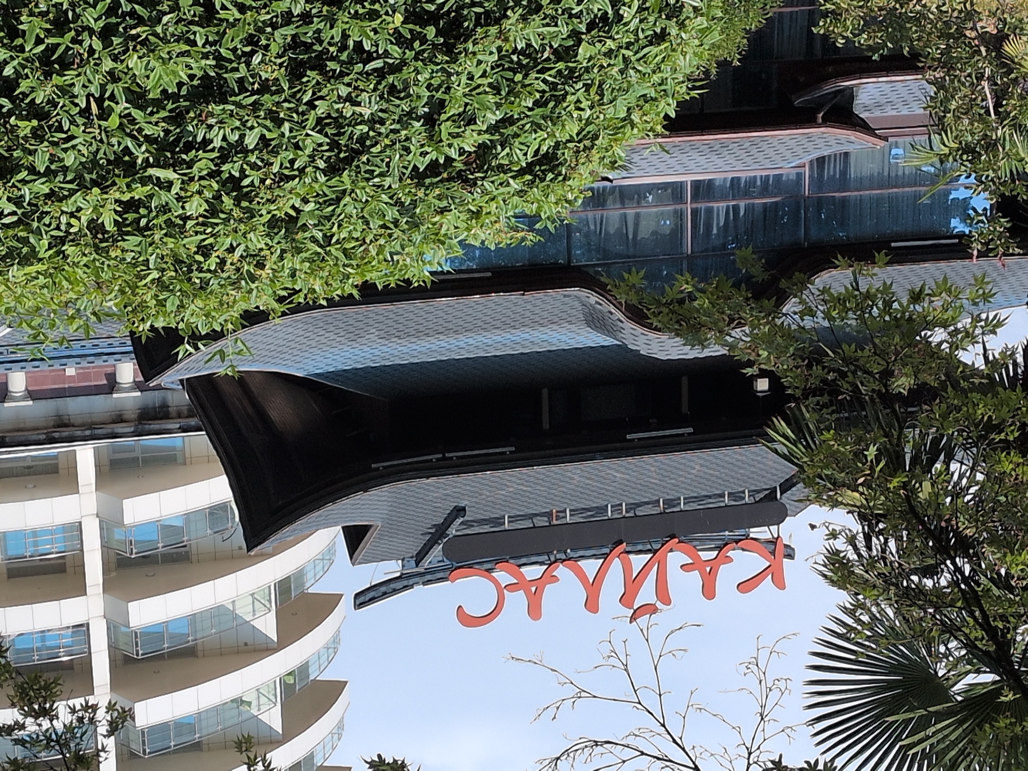 The image shows a building with a distinctive dark, multi-tiered, curved roof, reminiscent of traditional East Asian architecture. Red Cyrillic lettering spells out "КАЙЛАС" (Kaylas) across the top of the structure. This building is partially obscured by lush green foliage in the foreground and on the left side. In the background, a modern, white, multi-story building with curved balconies and glass railings stands against a pale sky. The overall scene suggests a location where modern and traditional architectural styles coexist, possibly a resort or a commercial establishment in a temperate climate, given the presence of greenery and the style of the buildings. This establishment is located in Guzeripl', Russia.