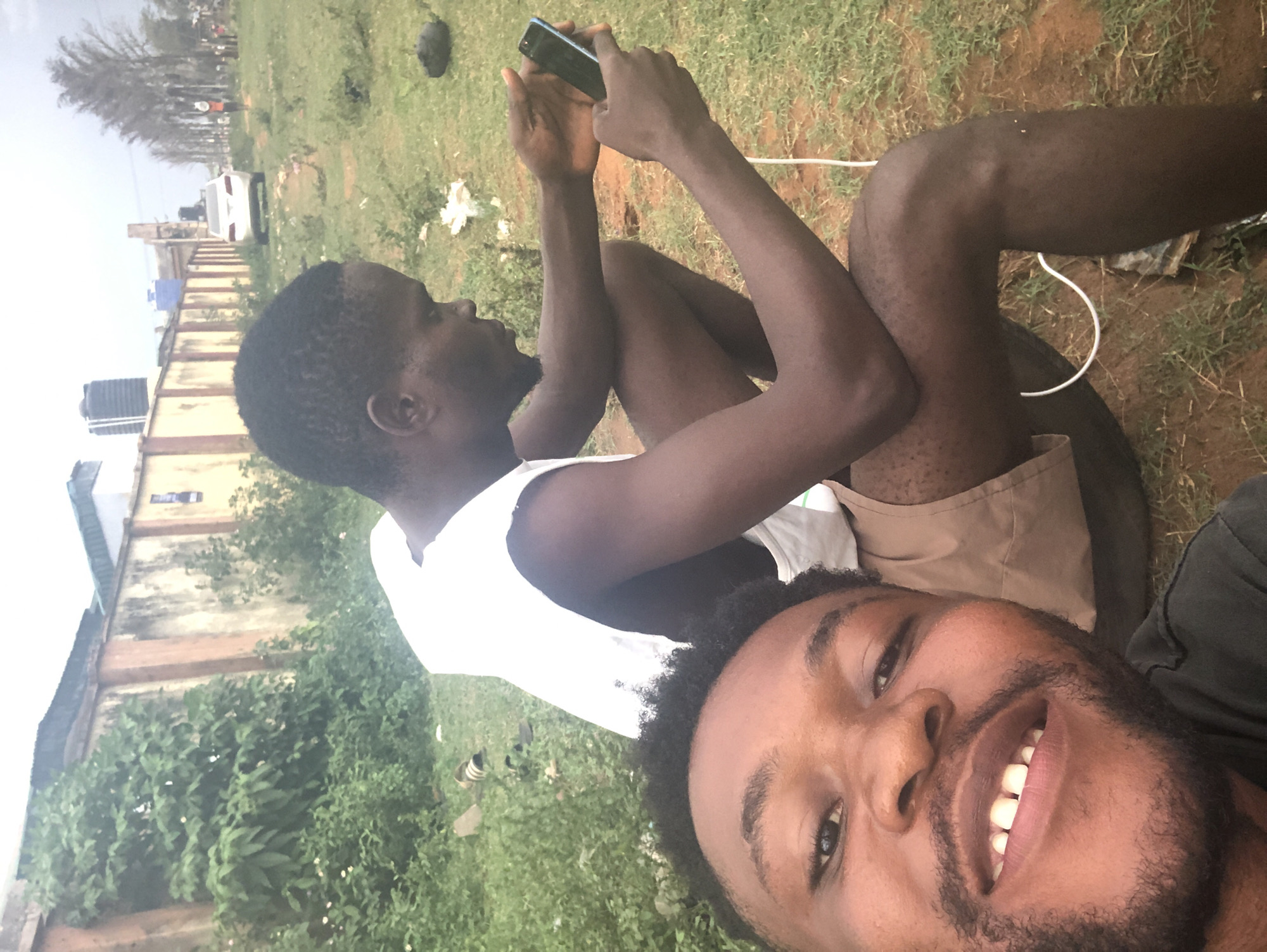 This media file captures a casual outdoor scene featuring two men. The image is a selfie taken by the man in the foreground, who occupies the lower right portion of the frame. He has dark skin, a short beard, and short dark hair, and is smiling broadly at the camera while wearing a dark-colored shirt.
Behind him, positioned to the left, another man is seated on the ground, engrossed in his smartphone. He wears a white tank top and light-colored shorts, with a white cable, possibly a charging cord, visible near his legs on the dirt.
The setting is an open, somewhat unkempt outdoor area characterized by patches of grass and exposed dirt. In the background, a long, low concrete wall separates the foreground from distant buildings. These structures, including one with blue and black water tanks on its roof, suggest an urban or suburban environment. A white car is visible parked in the distance behind the wall, and some sparse trees are seen against an overcast sky. The lighting indicates it is daytime, with diffused light rather than direct sunlight. The overall atmosphere appears relaxed and informal. No discernible text is visible.